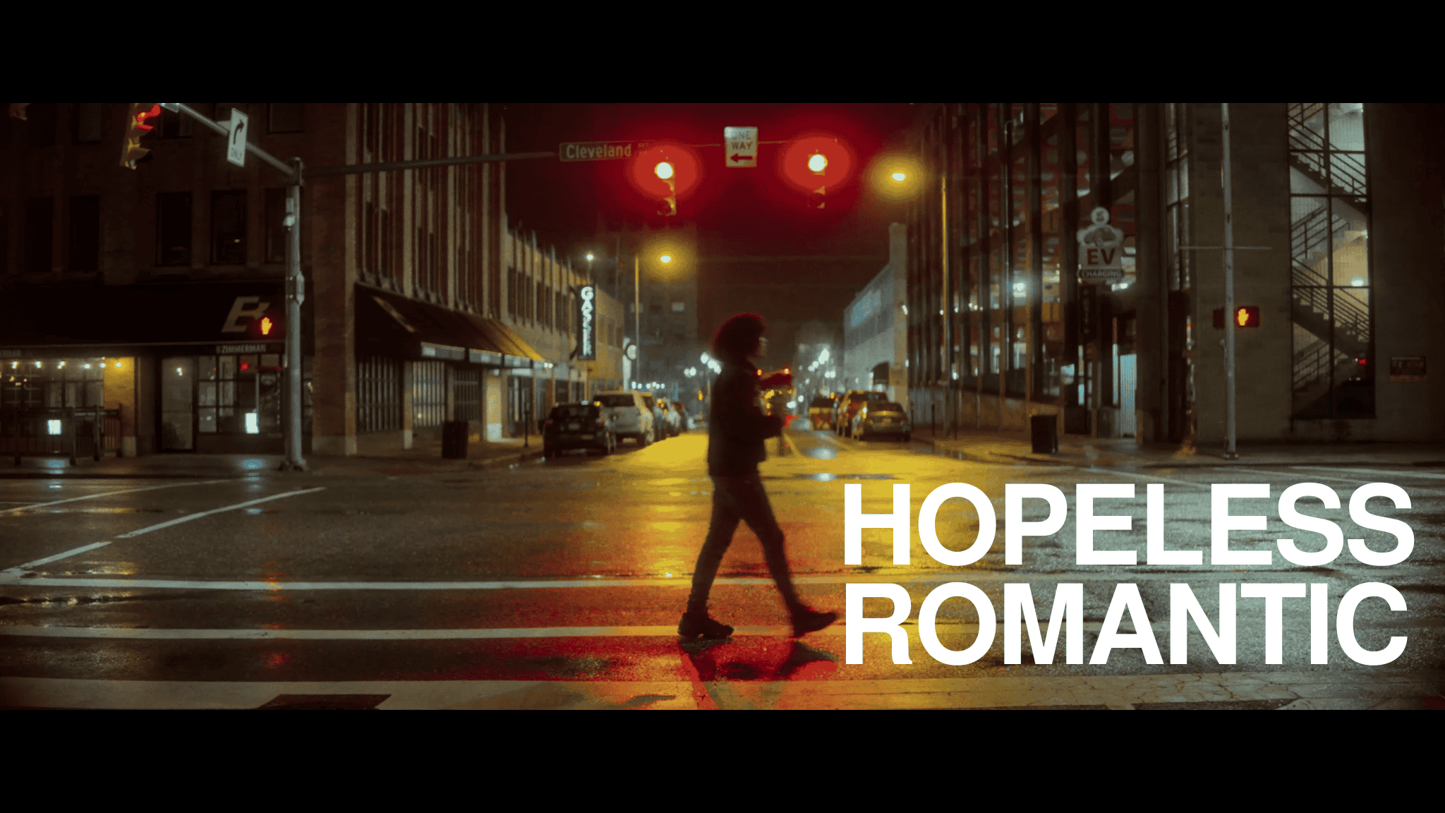 A person crossing a wet city street at night with traffic lights and parked cars, with the words "Hopeless Romantic" in white text.