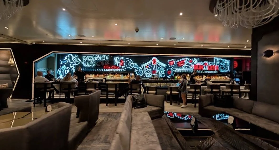 A modern bar with a neon sign display reading "Bright Lights Big City" behind the bar, which is set with bottles and glassware. There are patrons seated at the bar and couches in the foreground. The decor is sleek with dark tones and contemporary lighting.