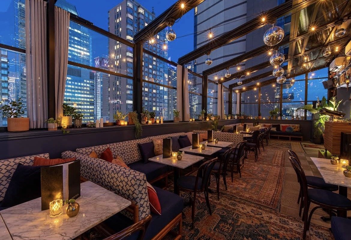 An indoor rooftop bar or restaurant with large glass windows overlooking a city skyline at dusk. The space is decorated with hanging string lights, candles, and potted plants. There are marble-top tables with chairs and a cushioned bench seating area.