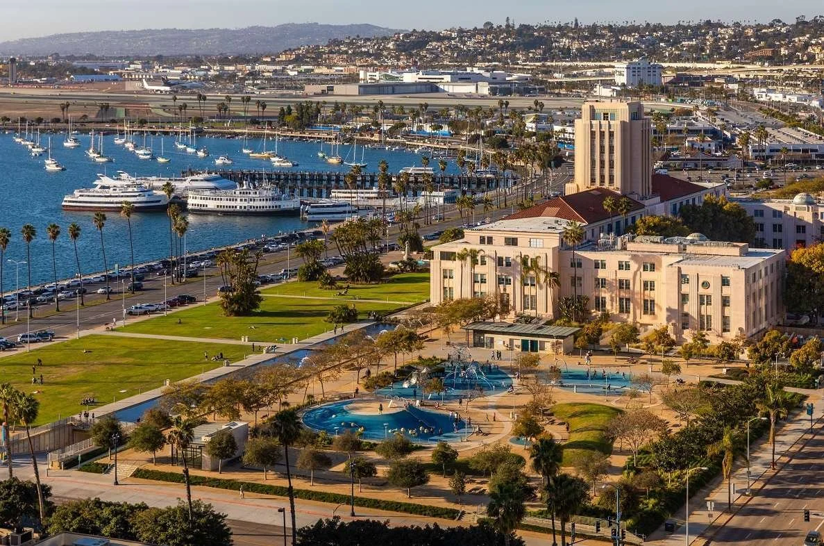 View of a city waterfront park with palm trees, water fountains, and people walking, in front of a marina with yachts and boats, and buildings and hills in the background.