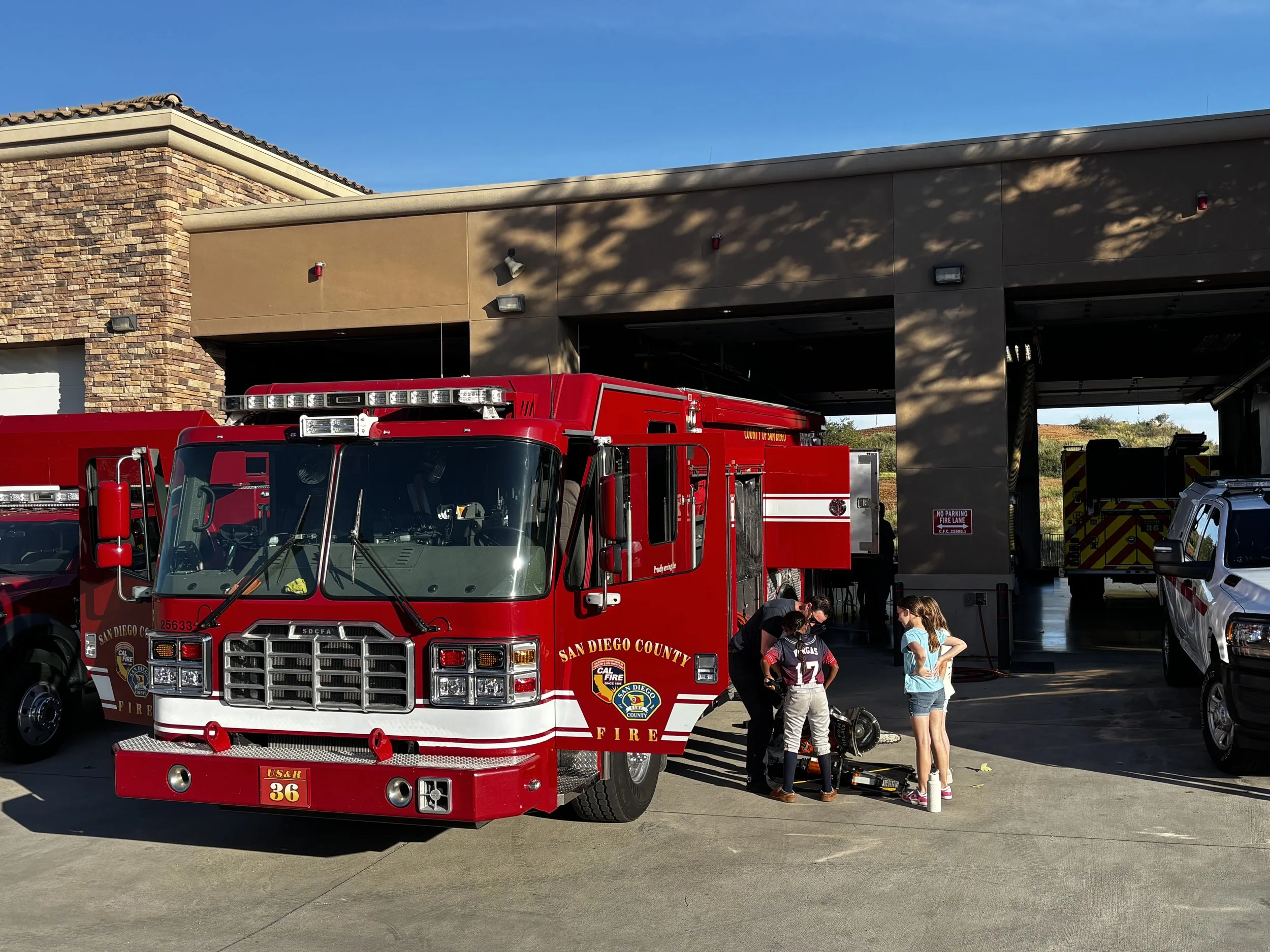 Multiple red fire trucks parked outside a fire station with children and adults standing nearby, under a sunny sky.