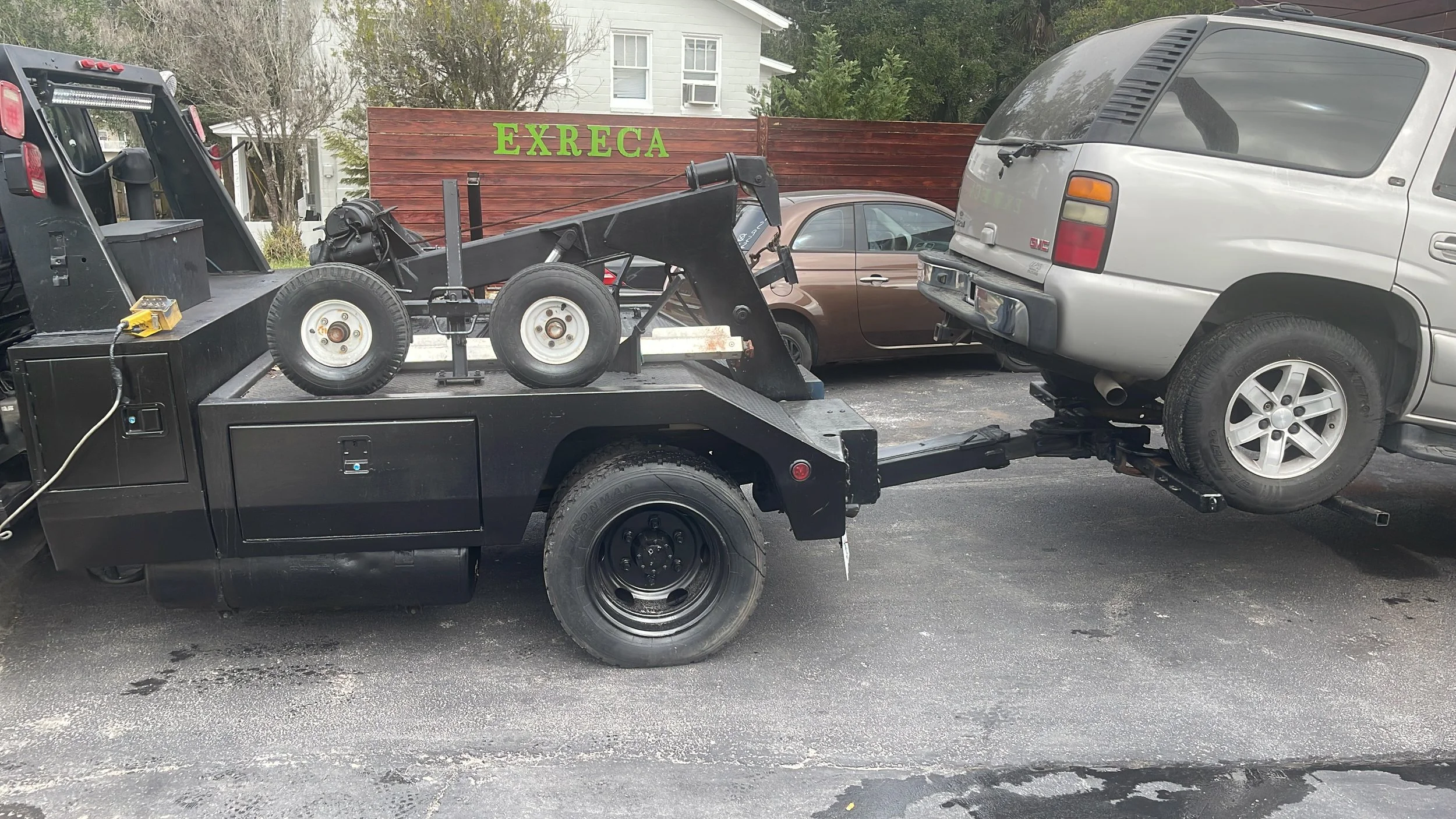 A flatbed tow truck with a black body is towing a silver SUV, which is partially elevated off the ground. In the background, other parked cars and a fence with a green 'EXRECA' sign are visible.