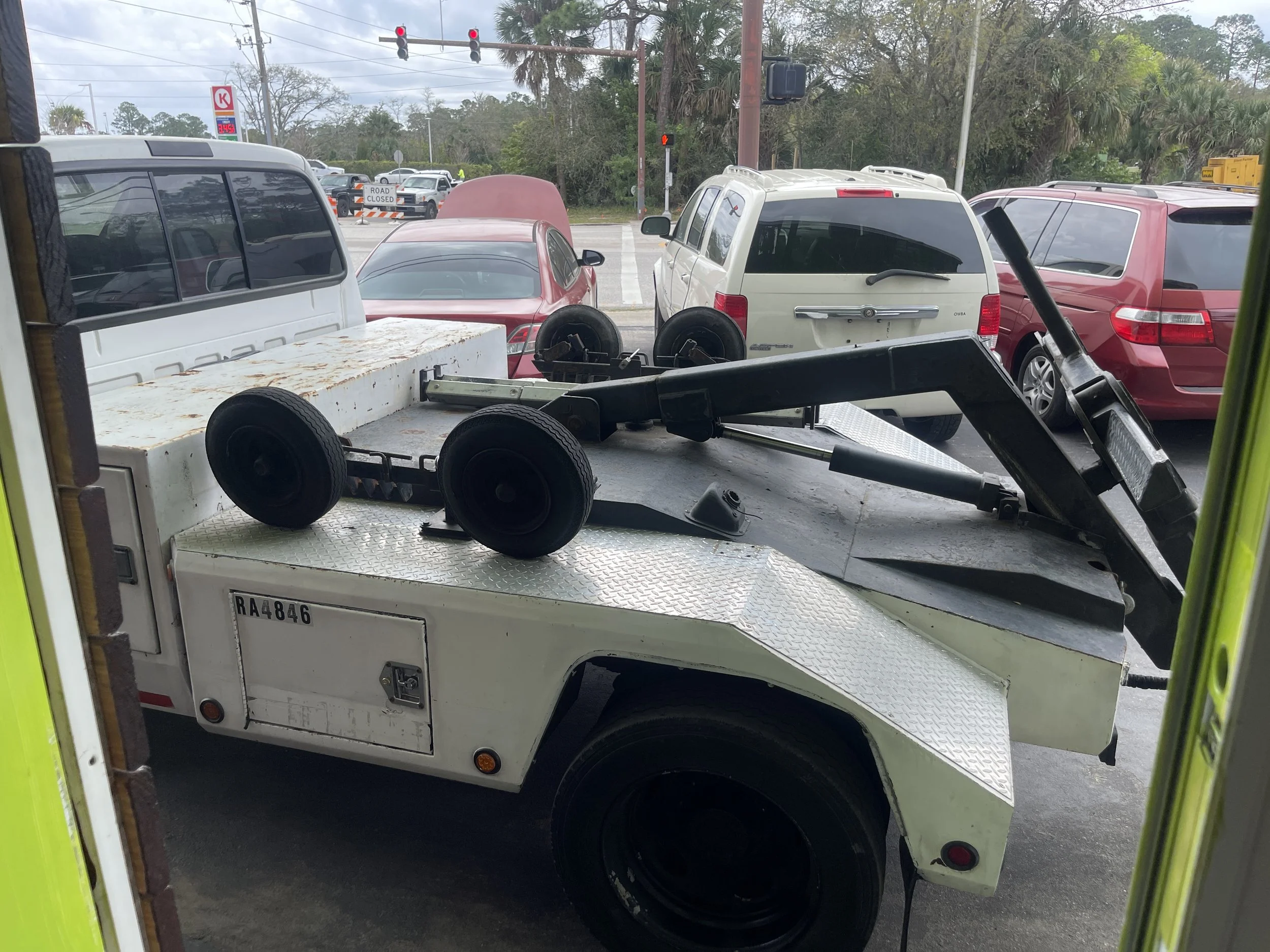 A flatbed tow truck with a metal platform, black wheel chocks, and control arm, parked in a parking lot with other vehicles and traffic lights visible in the background.