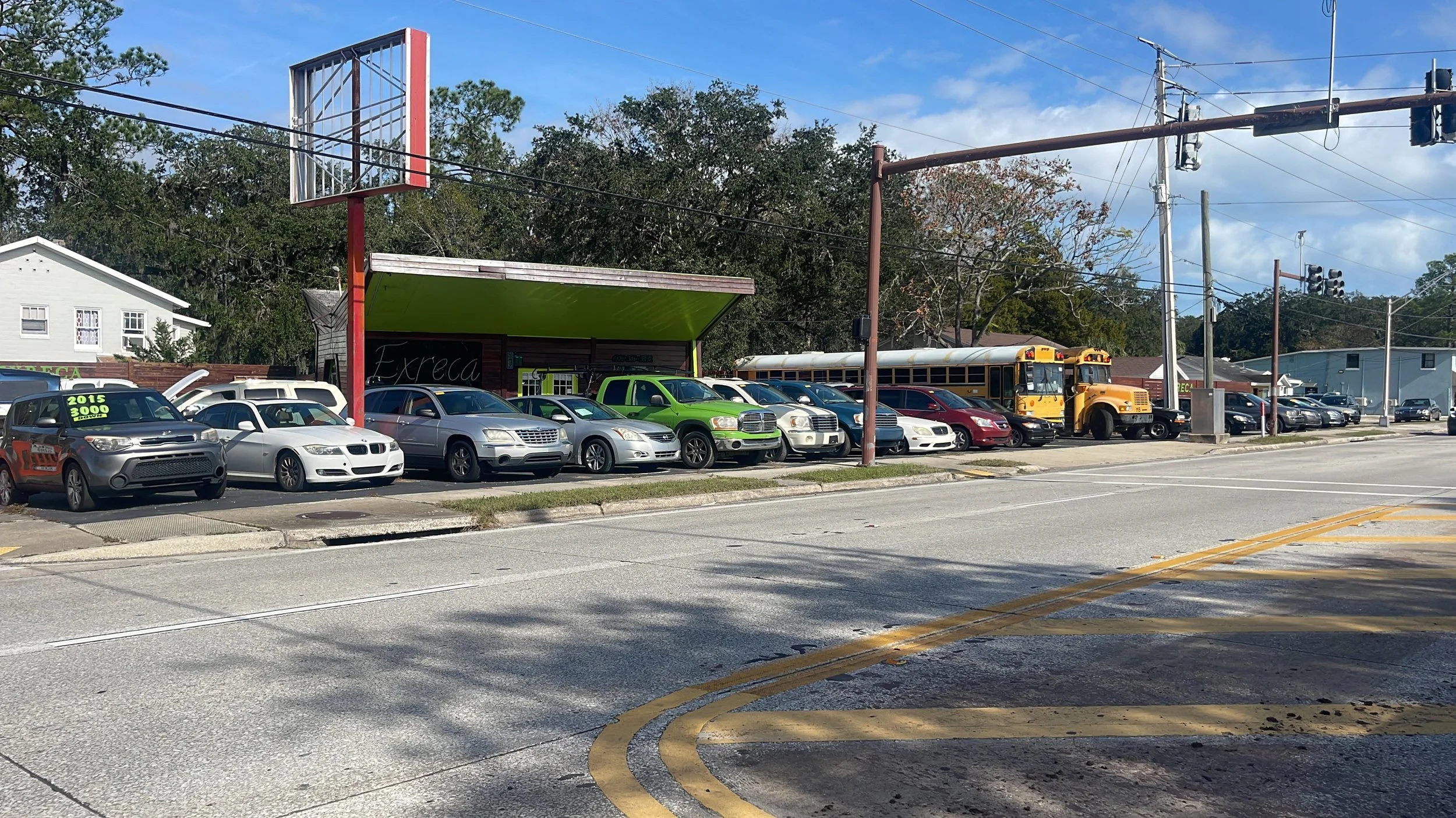 A parking lot with various cars including a gray sedan, a white convertible, a silver minivan, a green truck, and a school bus, with a sidewalk and street in the foreground and power lines overhead.