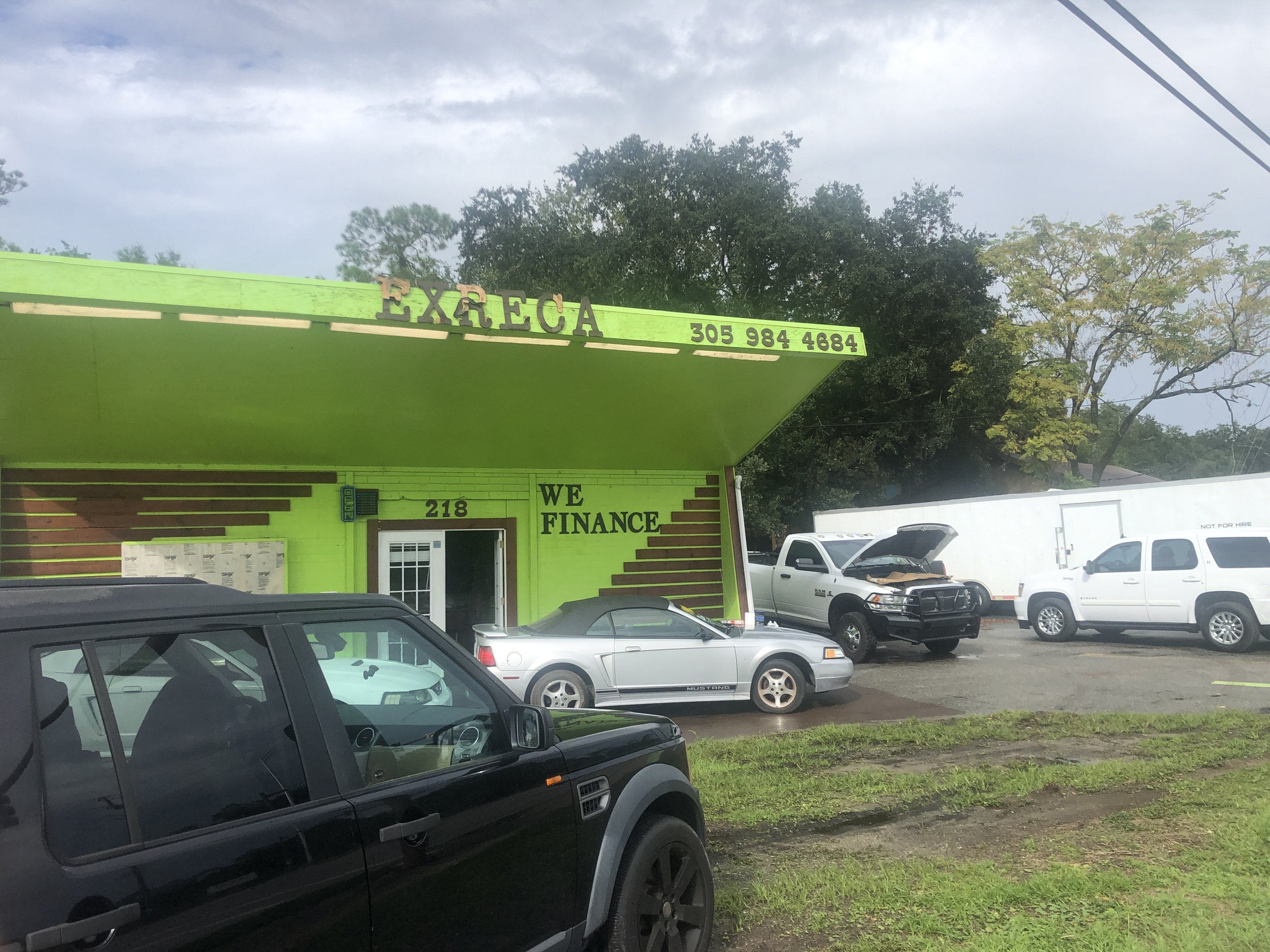 A colorful business storefront painted in bright lime green with the sign 'EXRECA' and phone number 305 984 4684. Several cars parked in front, including a black SUV, a silver convertible, and white trucks, with some vehicles having open hoods. Tall trees and partly cloudy sky in the background.