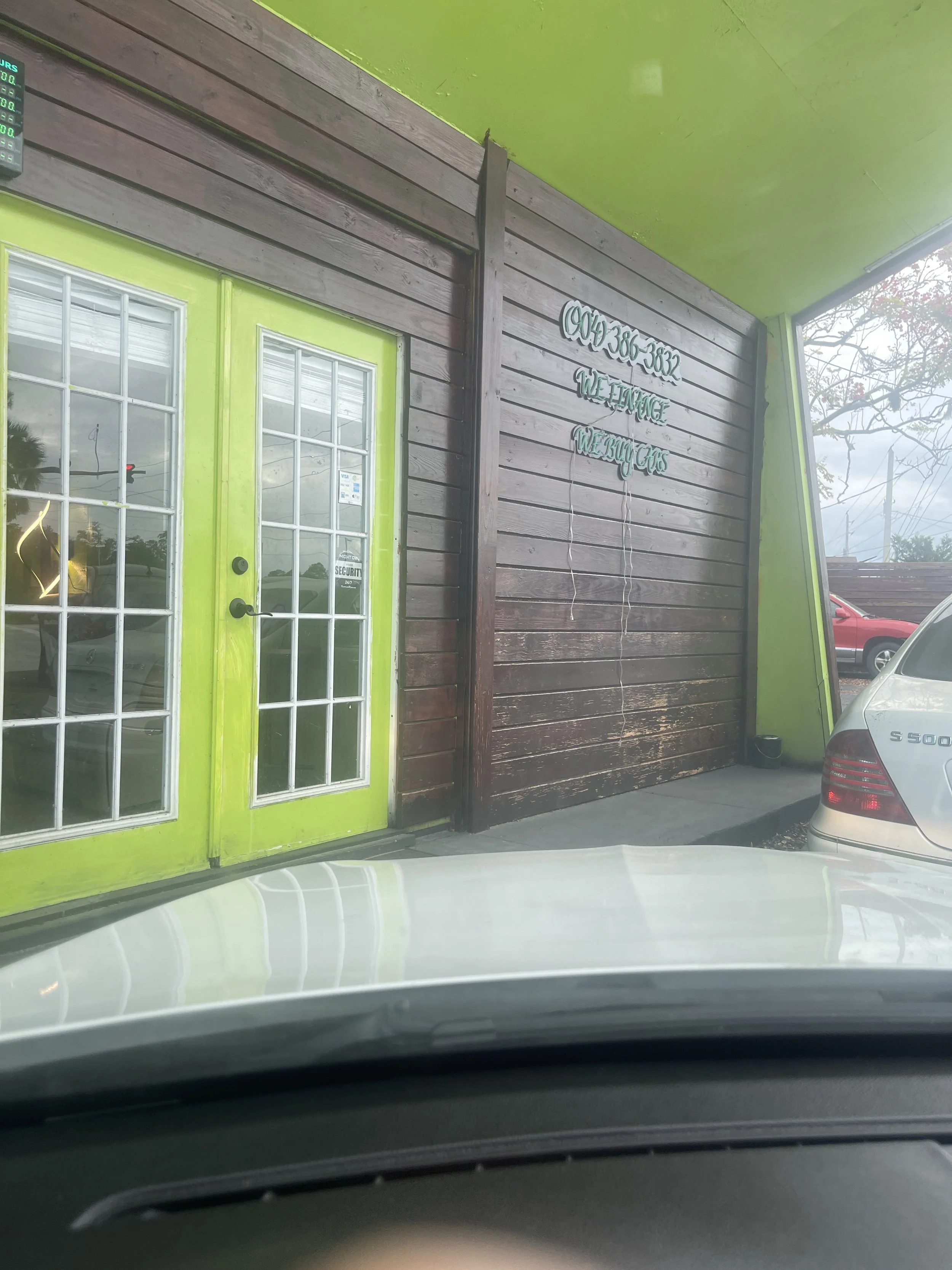 View of the front entrance of a cannabis dispensary with bright green doors and a dark wood-paneled wall. Signs display the phone number and slogan 'We iNjoy, We BuY, and We iNvolve.' There is a parked silver Mercedes-Benz S-Class and vice versa, the photo is taken from inside a car.