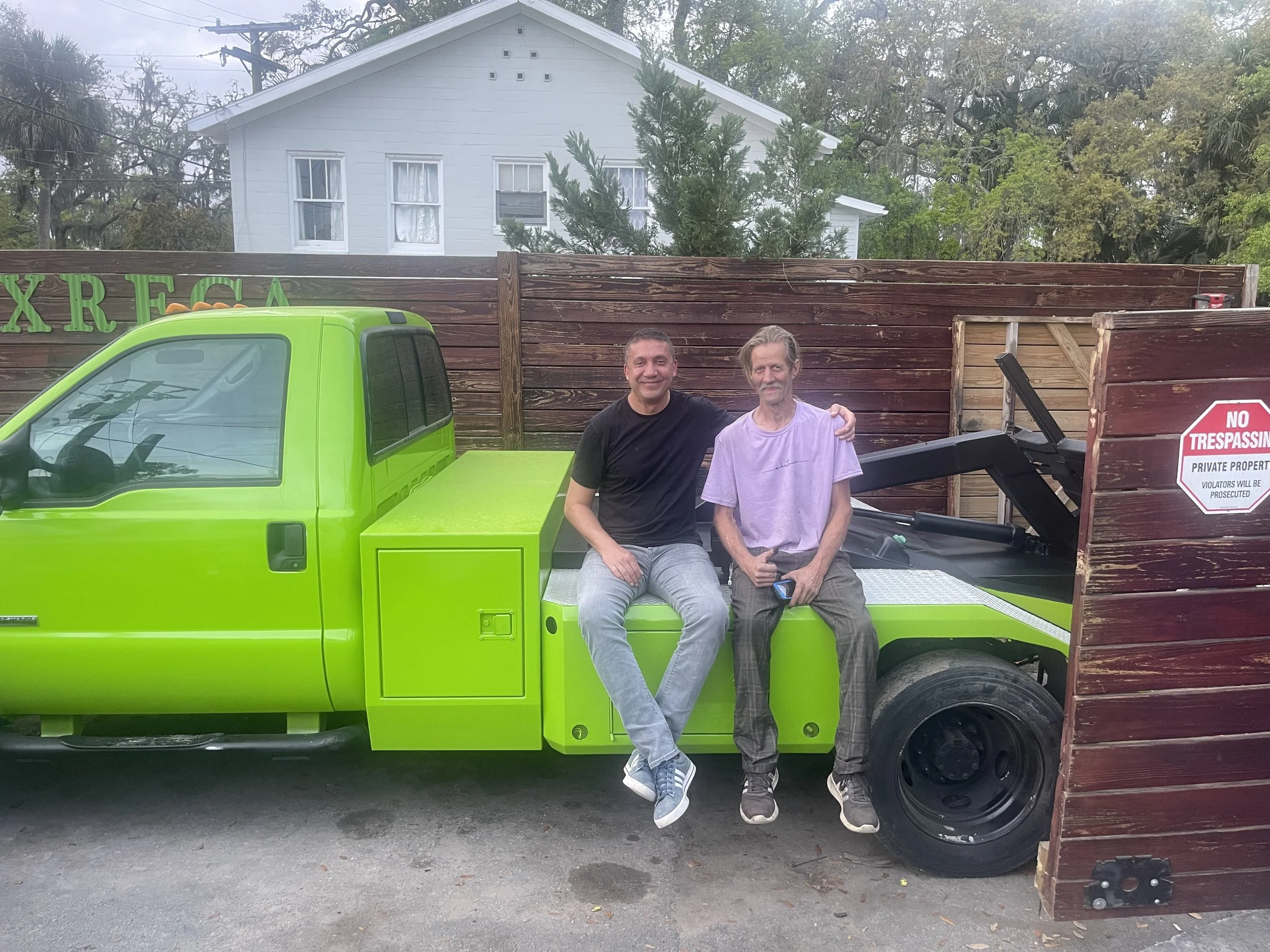 Two men are sitting on the side of a bright green Tow truck parked in front of a wooden fence. One man is wearing a black T-shirt and gray jeans, and the other is wearing a light purple T-shirt and plaid pants. There is a red and white 'No Trespassing' sign on the fence.