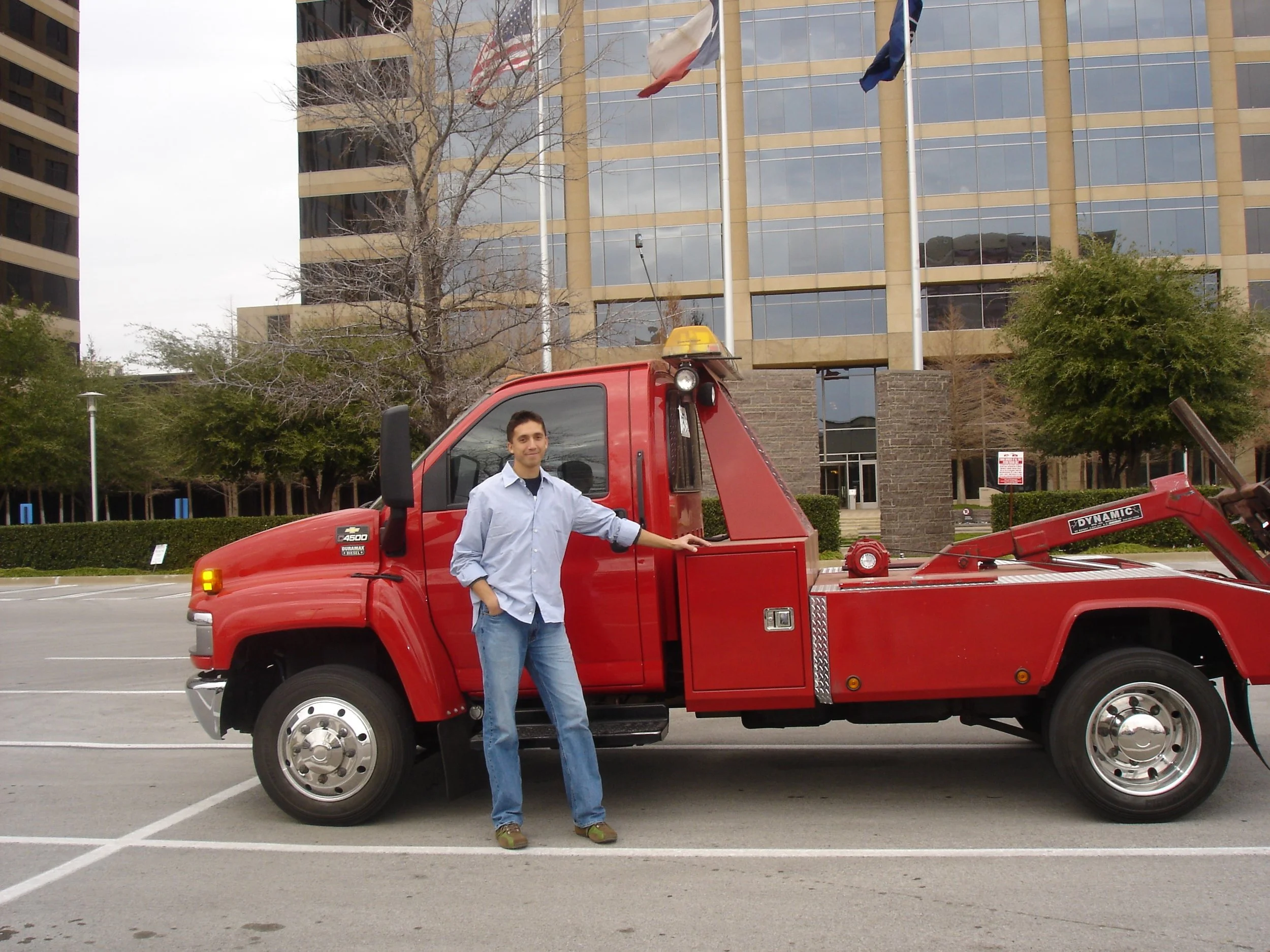 A man is standing next to a red tow truck in a parking lot. There is a large building with glass windows and flags in the background, along with some trees and a cloudy sky.