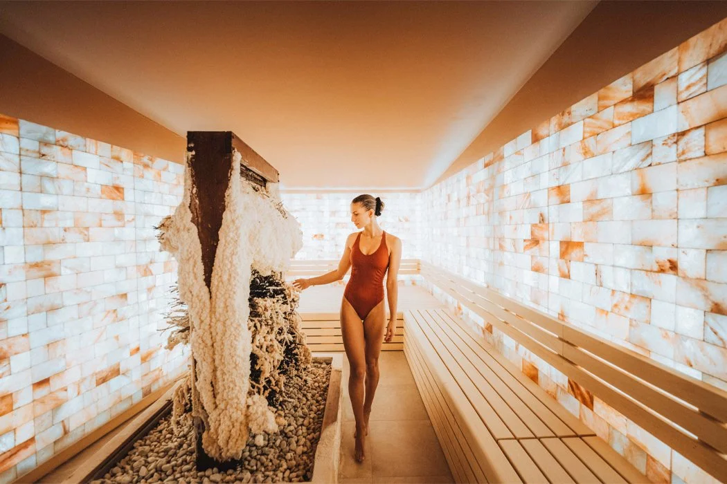 A woman in a red swimsuit and black hair tied back is walking in a spa or sauna area, touching a stone wall in a room with warm lighting, salt-infused walls, and a wooden bench.