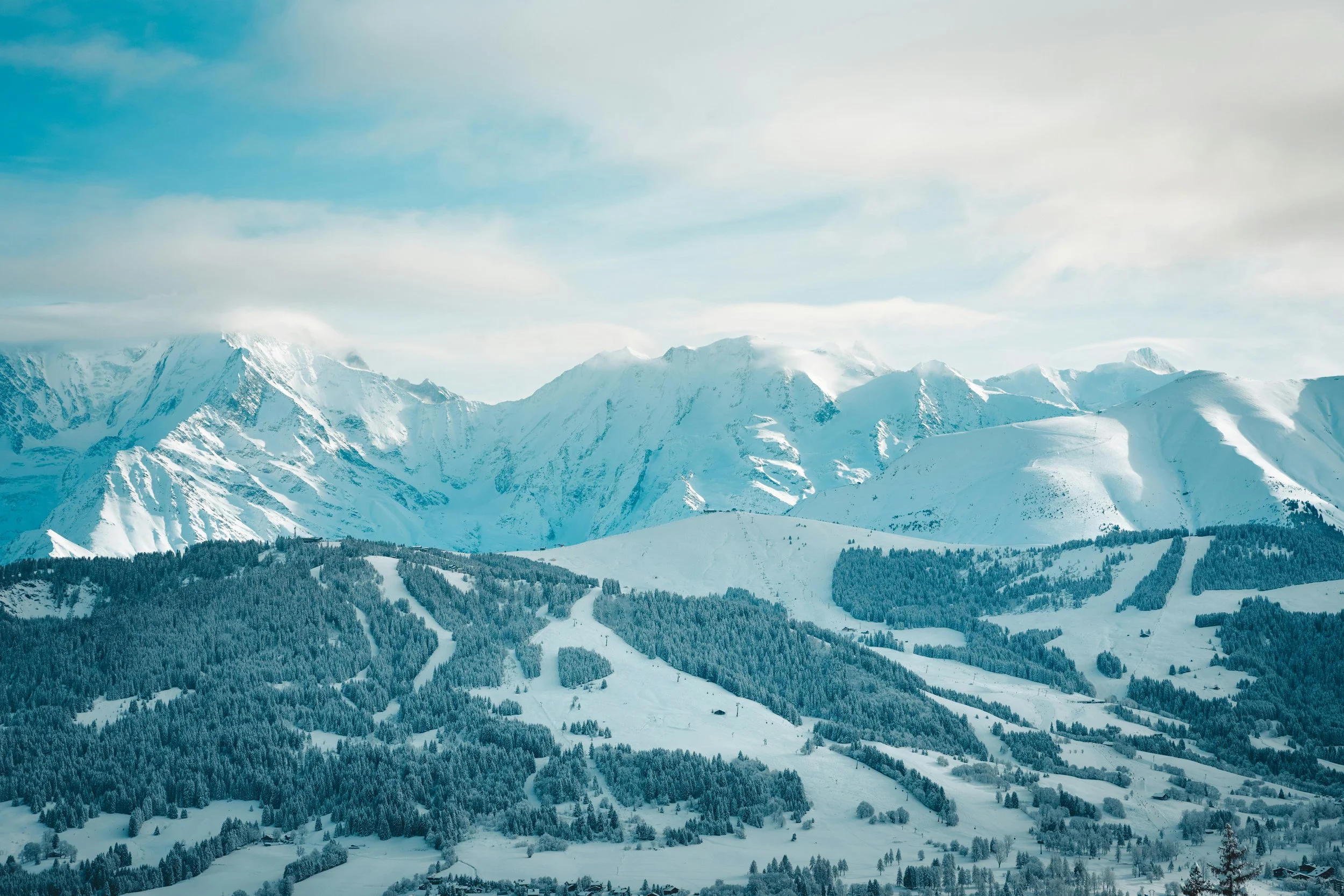 Snow-covered mountain range with pine forests and ski slopes under a partly cloudy sky.
