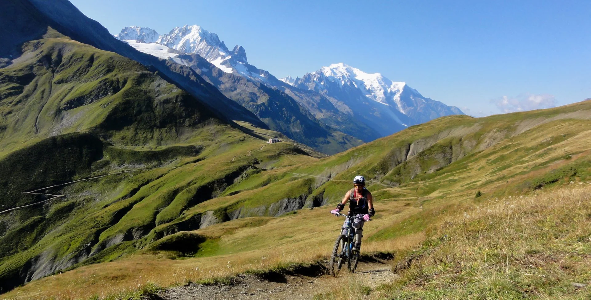 A woman mountain biking on a trail through a scenic green valley with snow-capped mountains in the background under a clear blue sky.