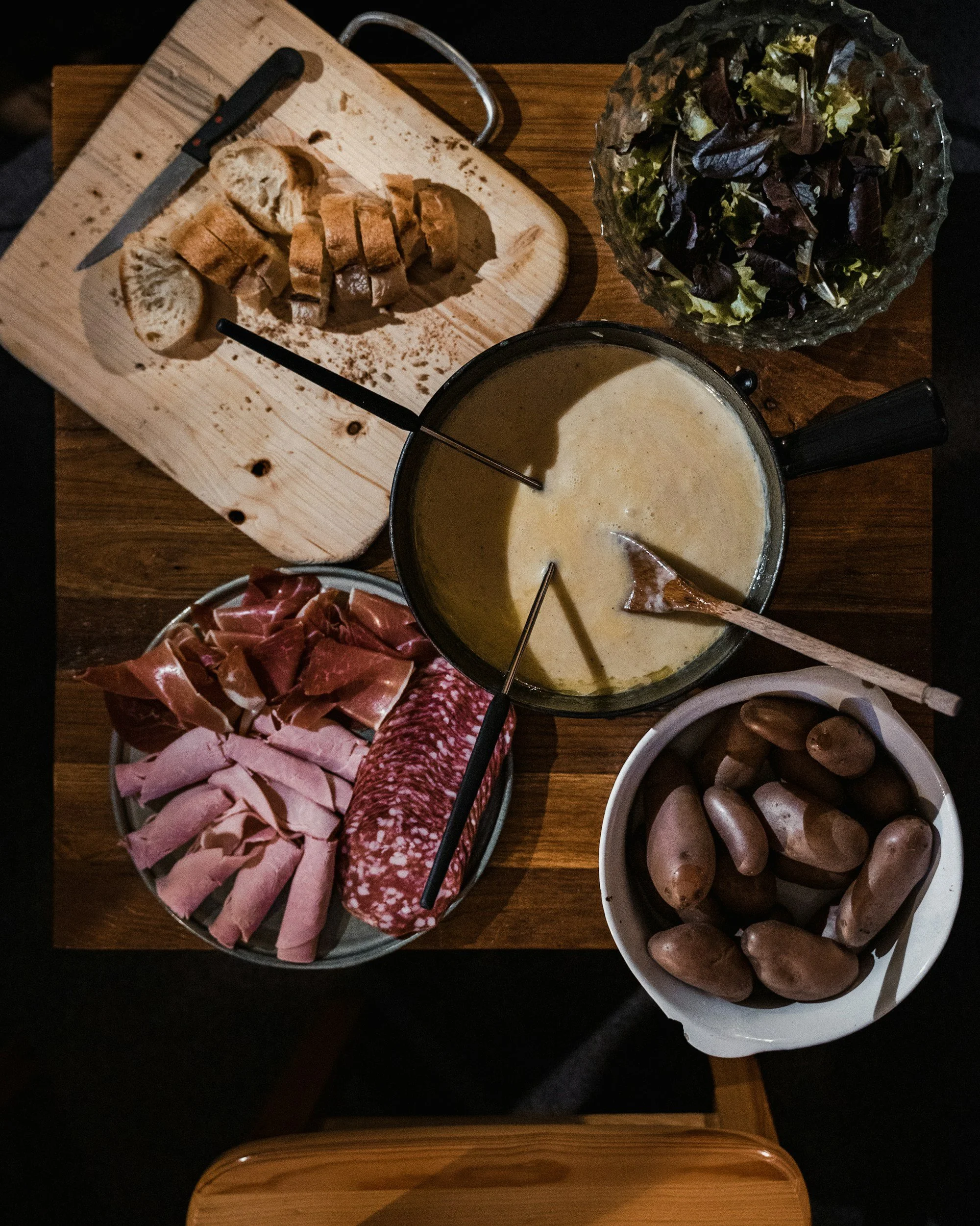 A cheese fondue setup with a pot of melted cheese, a bowl of assorted meats including prosciutto, salami, and turkey slices, a bowl of brown mushrooms, a bowl of mixed salad greens, and a wooden cutting board with sliced bread and a knife, all on a w