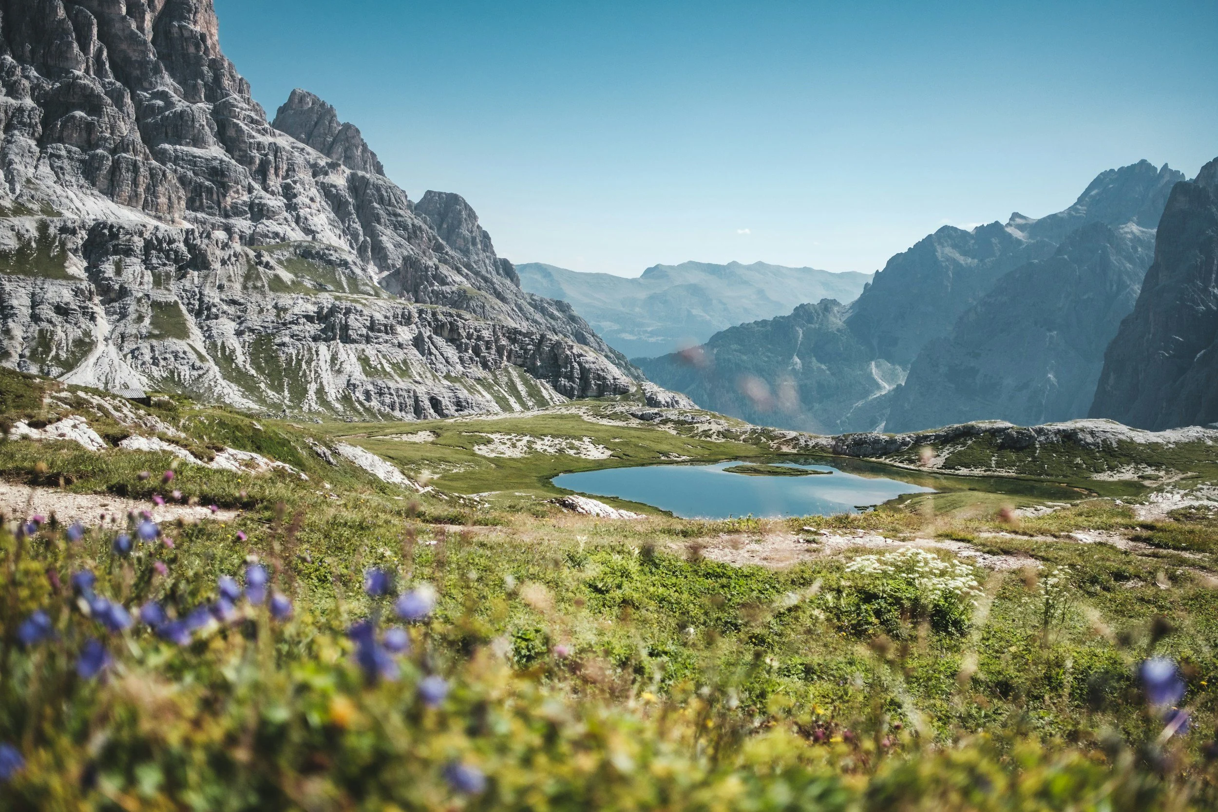 Scenic mountain landscape with rocky peaks, a small lake, and green meadows with wildflowers under a clear blue sky.
