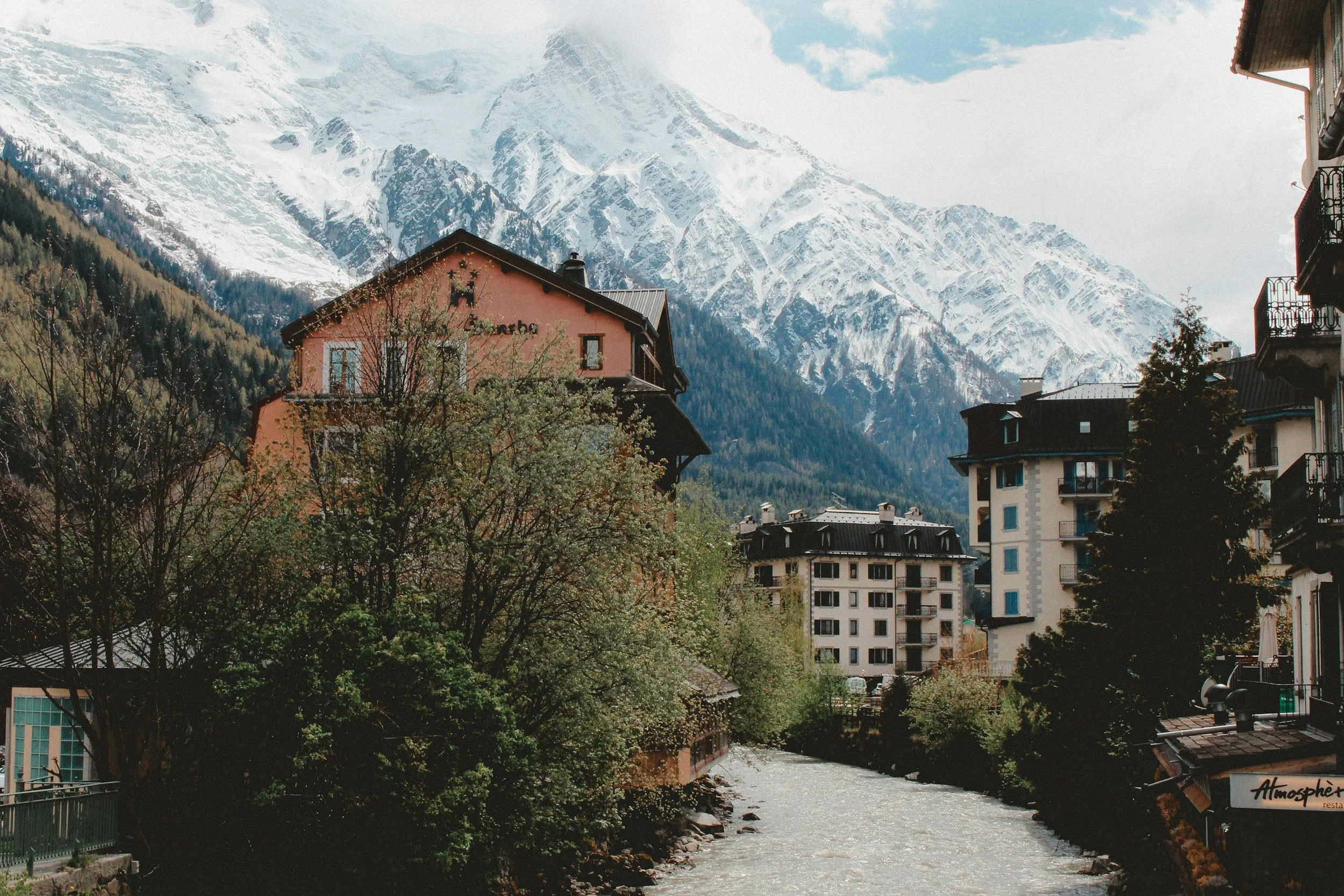 A river flows through a small town with colorful buildings, trees, and snow-capped mountains in the background.