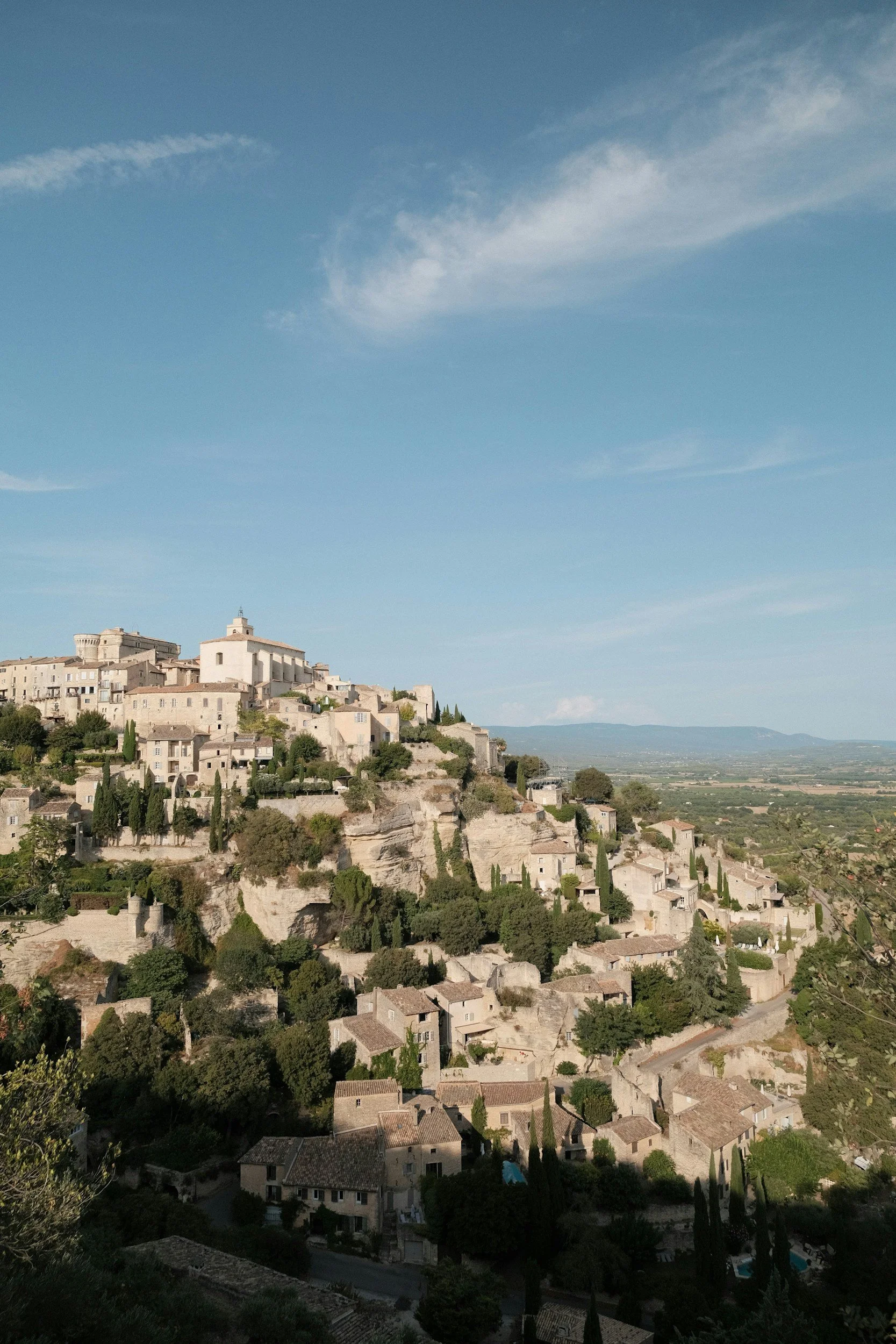 A hillside village with stone houses and cypress trees under a blue sky with wispy clouds.