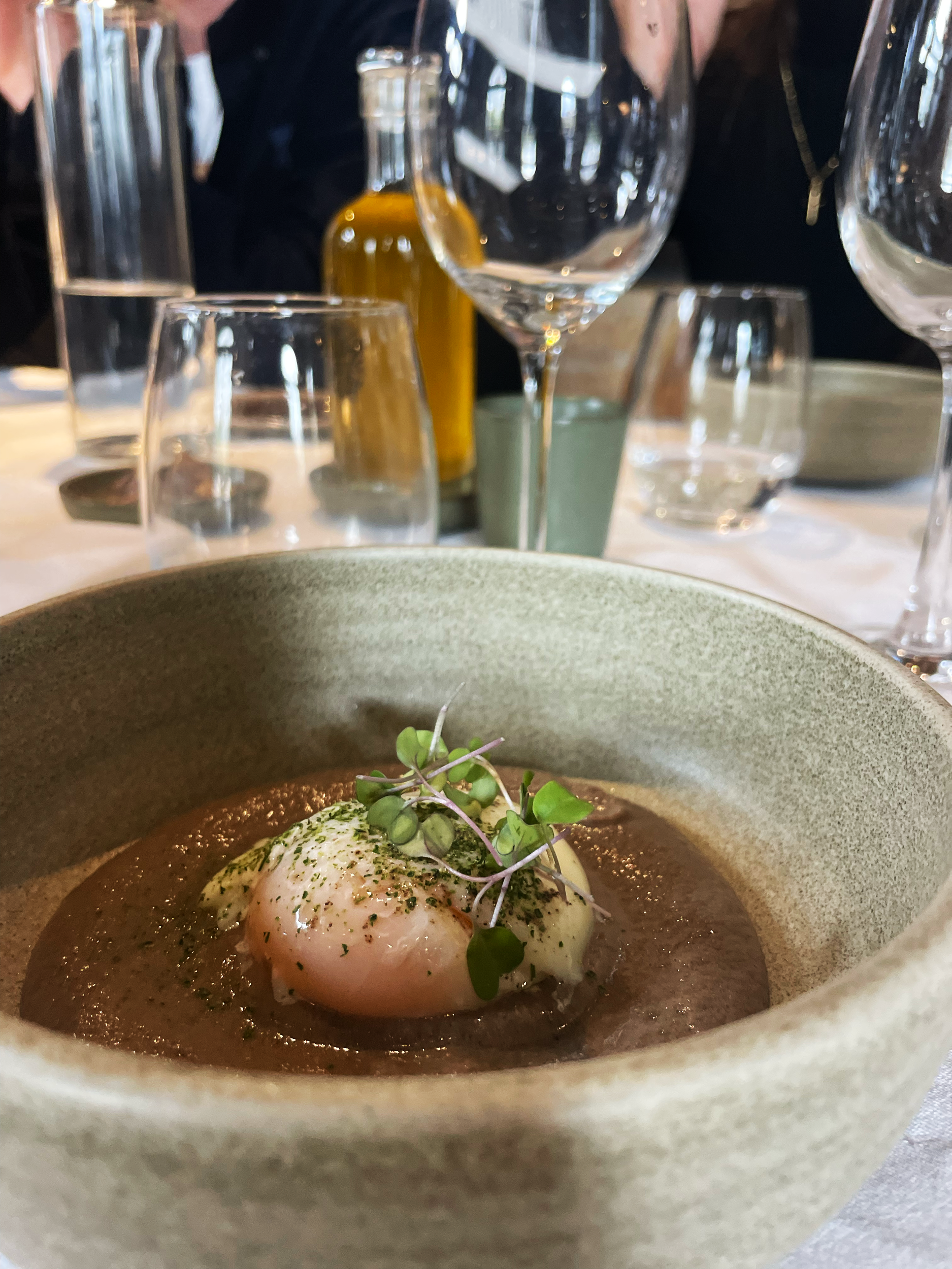 A close-up of a bowl with poached eggs garnished with microgreens on a table at a restaurant, with wine glasses, water glasses, a bottle of olive oil, and other tableware in the background.