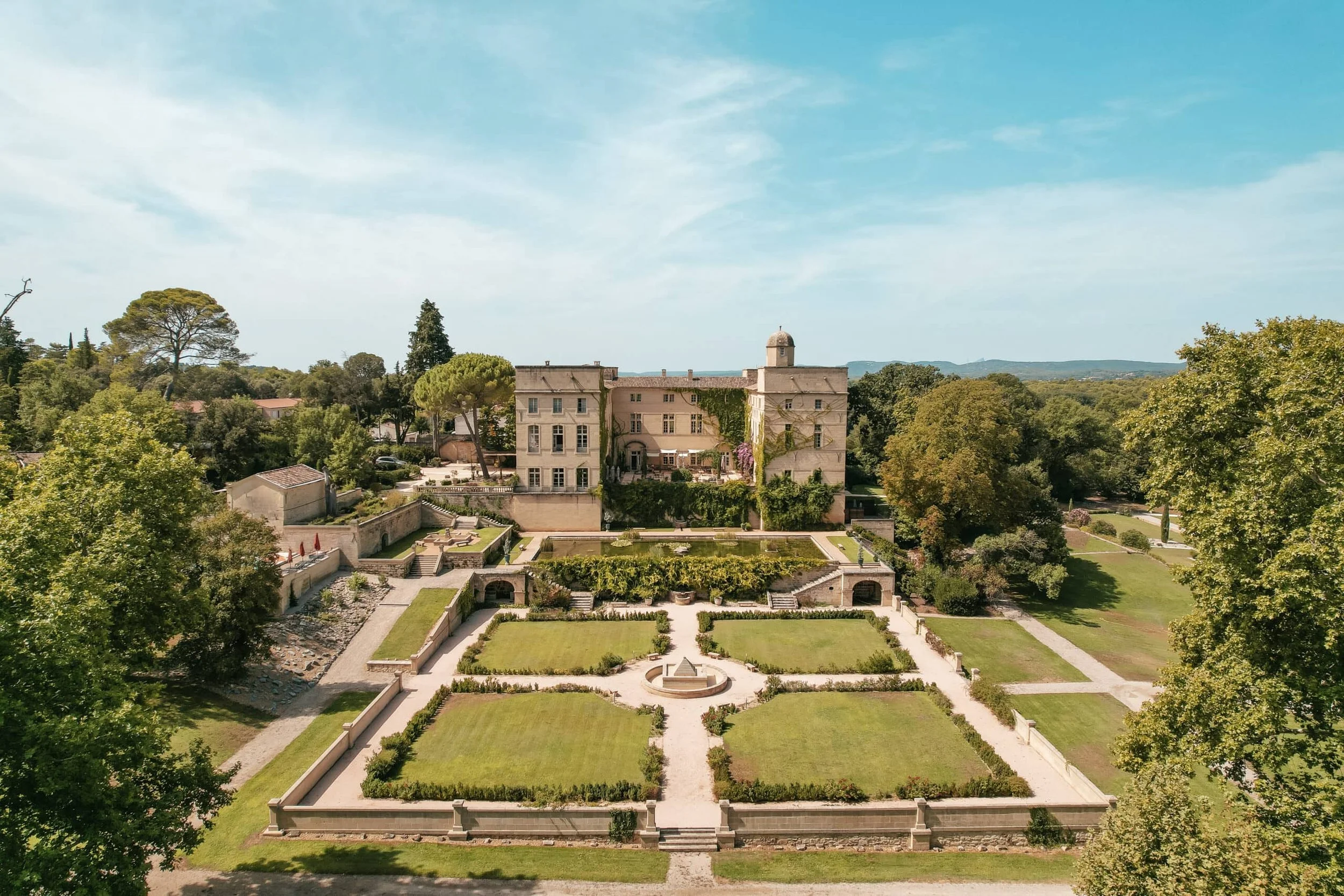 A large historic mansion with a garden in front, surrounded by tall trees under a clear blue sky.