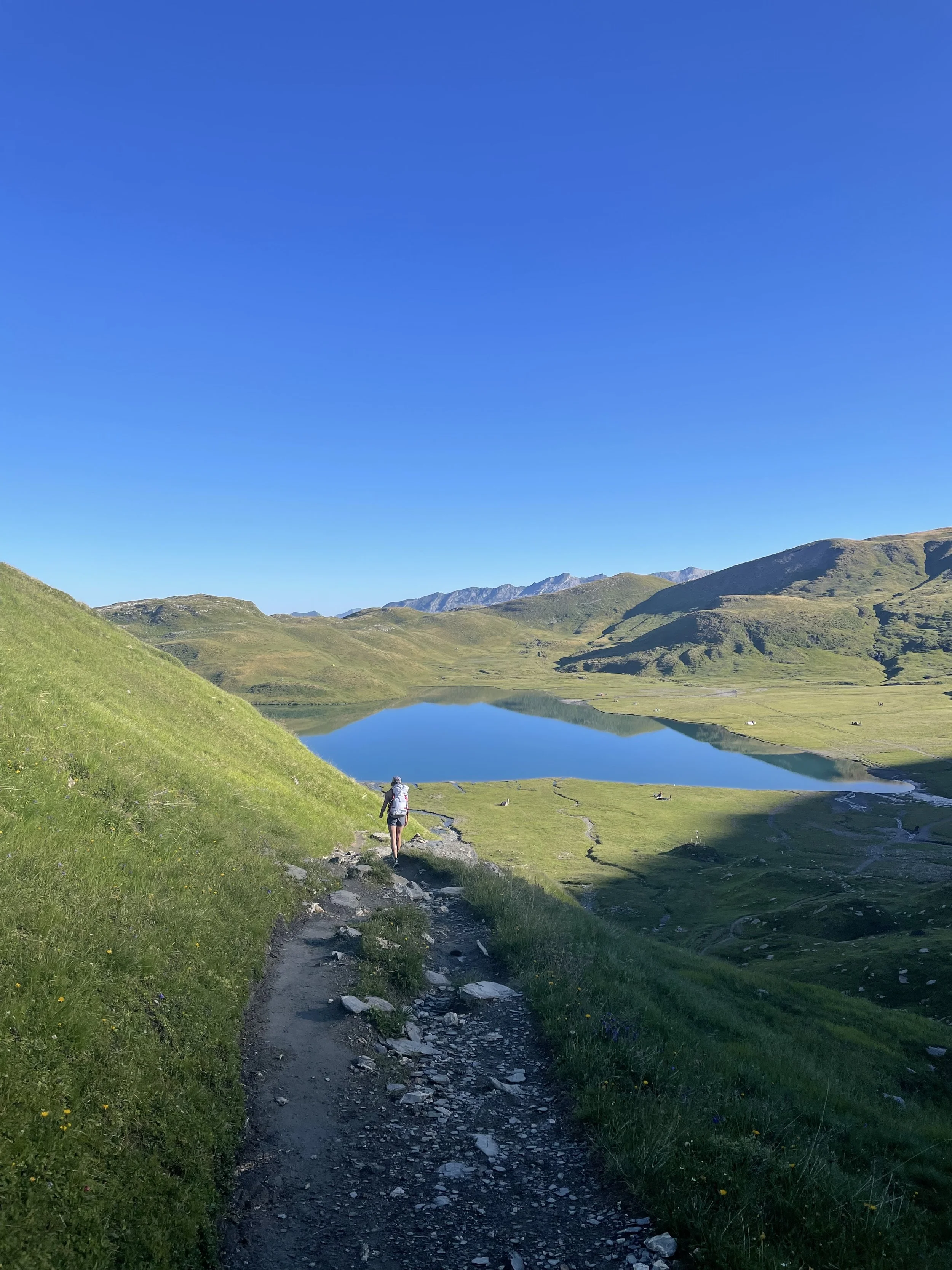 Hiker walking on a narrow dirt trail through green grass hills towards a calm lake surrounded by mountains under a clear blue sky.