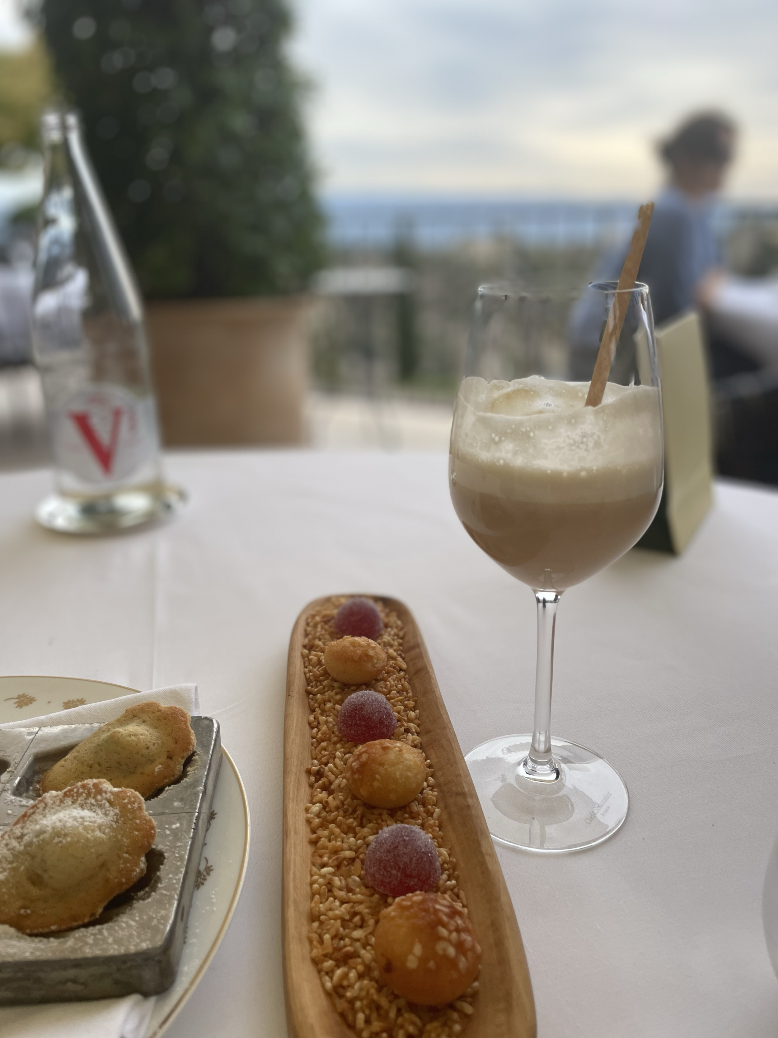 A table set with a glass of iced coffee with whipped cream and a stir stick, a long wooden platter with small bites topped with colorful toppings, a metal tray with pastries, and a glass bottle of water, with a blurred outdoor background including a
