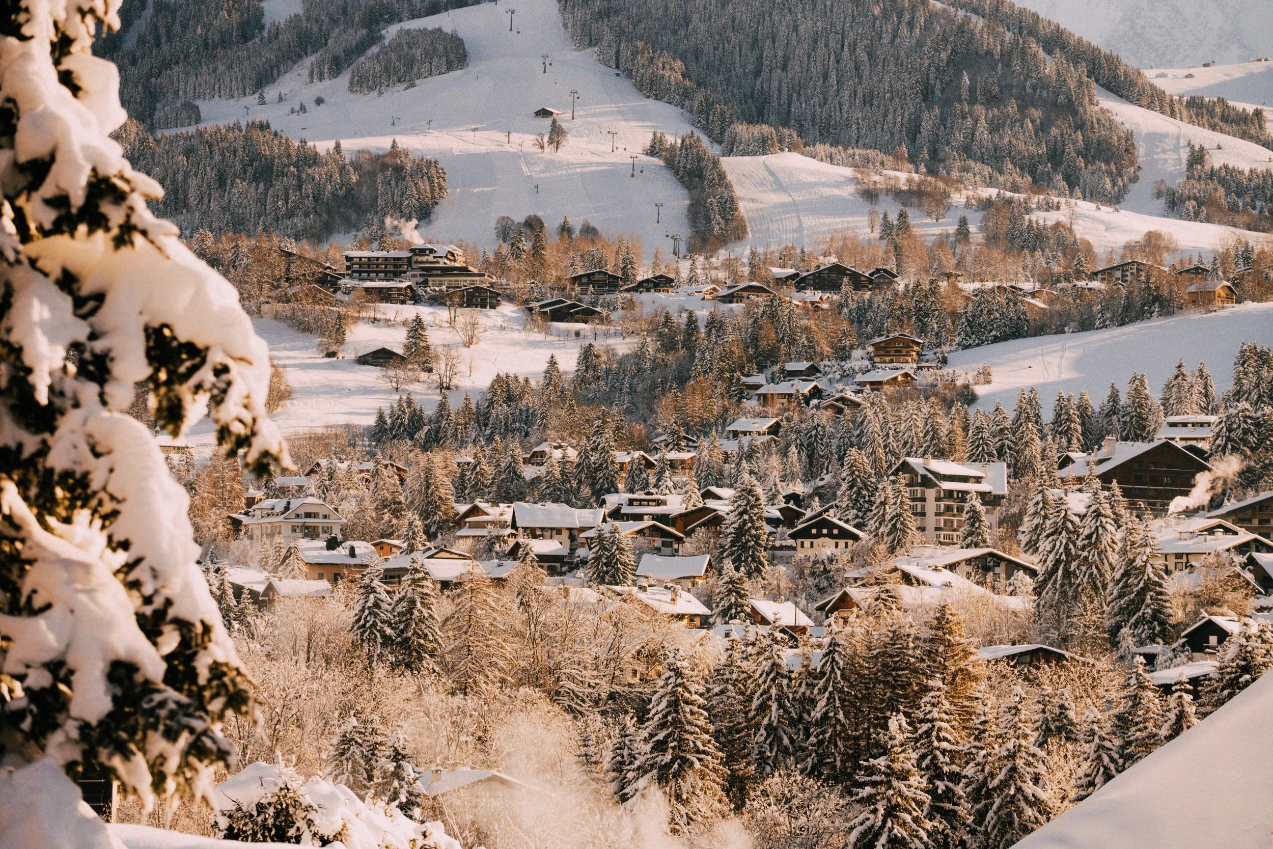 Snow-covered village nestled in a forested mountain landscape during winter at sunrise.