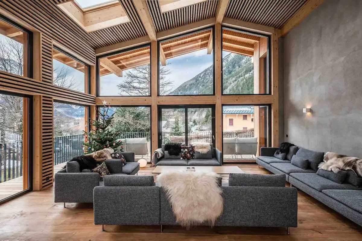 A modern living room with large floor-to-ceiling windows showing a mountain view, decorated for Christmas with a Christmas tree and plush throws on gray sofas.