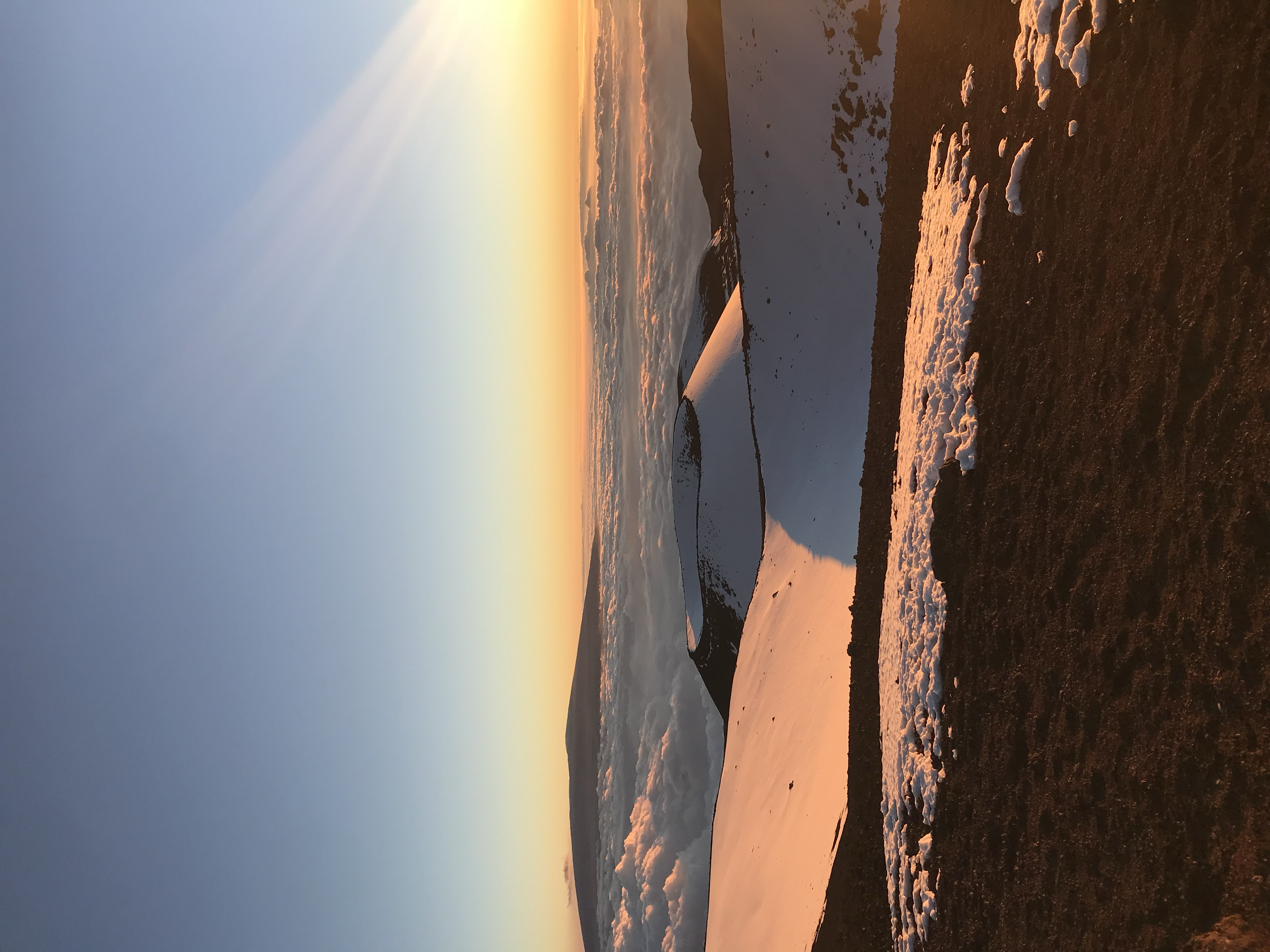 Snow-covered mountain peaks at sunset with the sky glowing in warm orange and pink hues, and a barren landscape in the foreground.