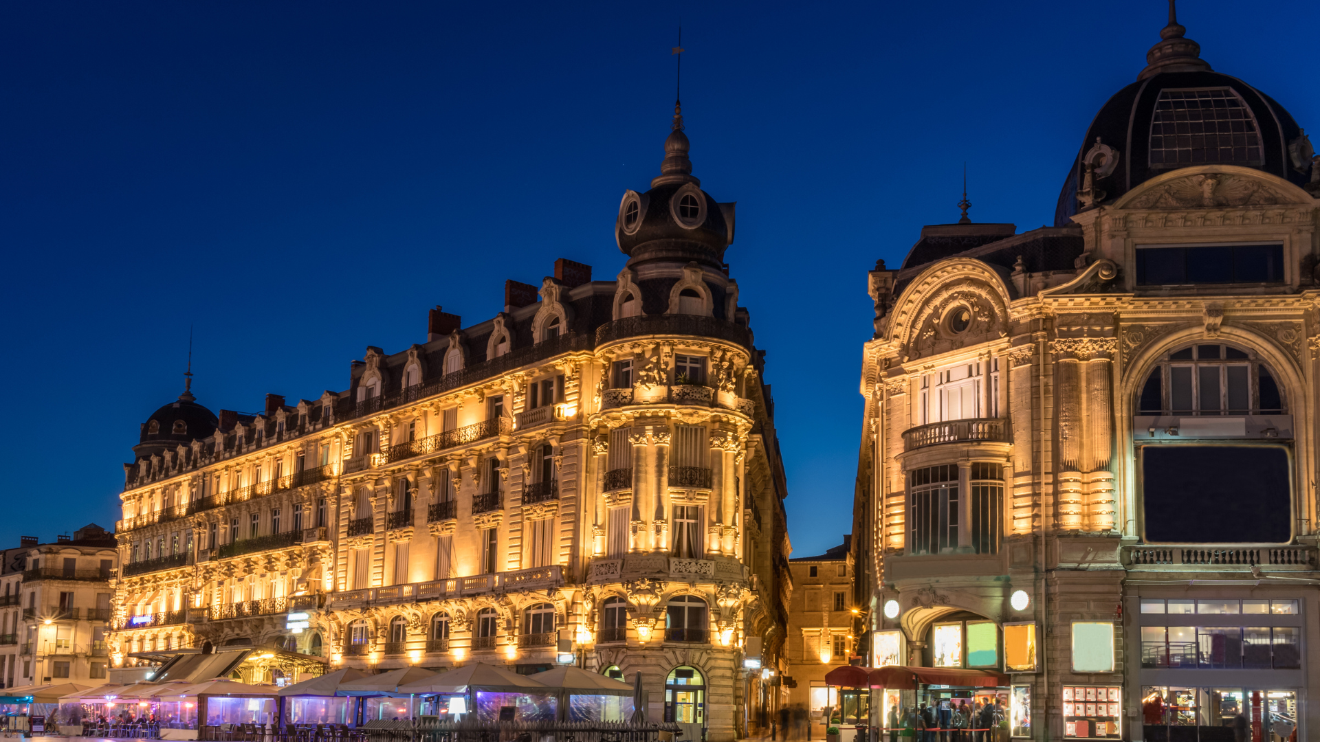 Illuminated historic buildings with ornate architecture at dusk, cafes with outdoor seating, and a clear blue sky.