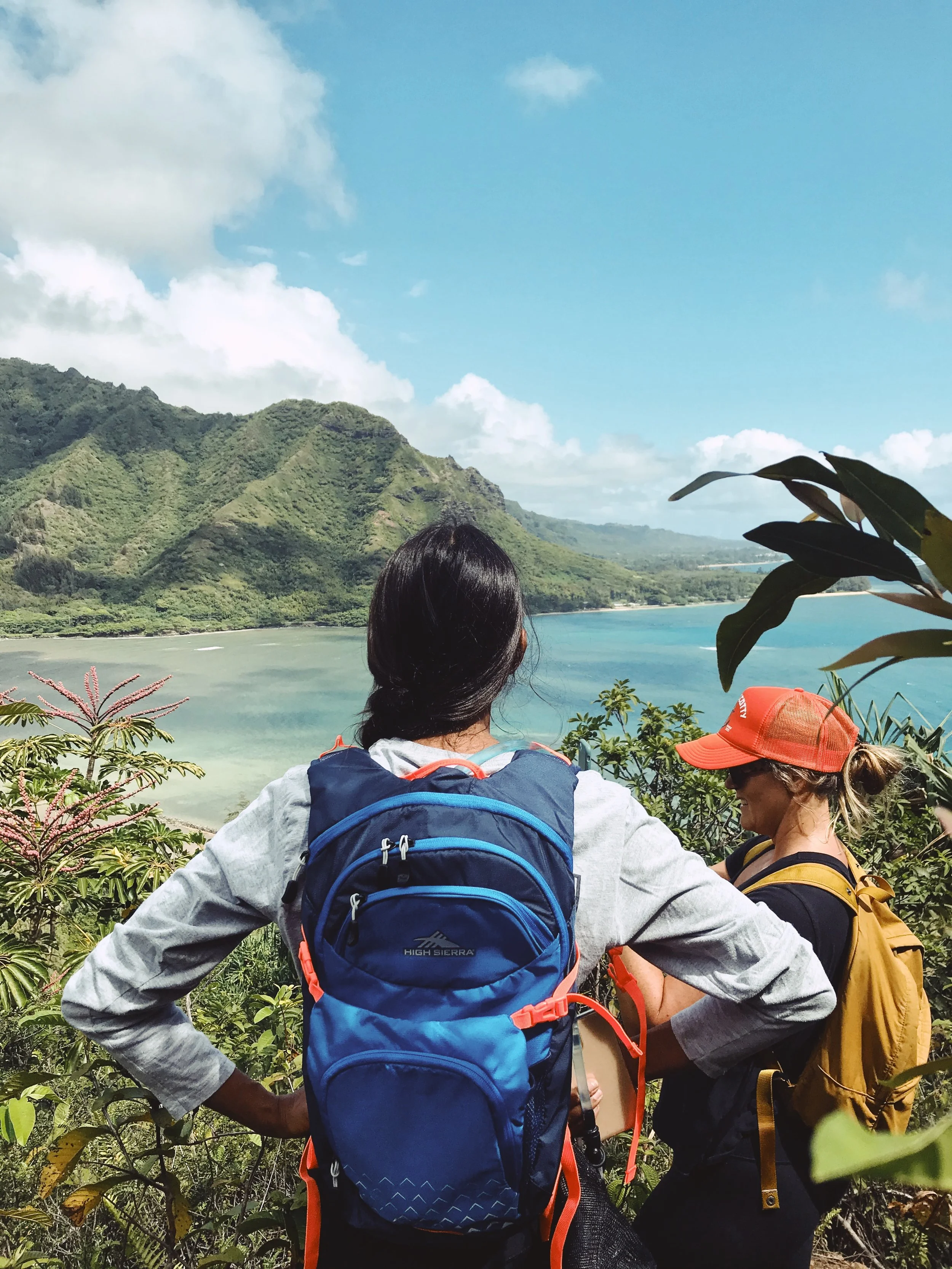 Two women hiking with backpacks overlook a scenic view of mountains, a lake, and lush greenery under a partly cloudy sky.