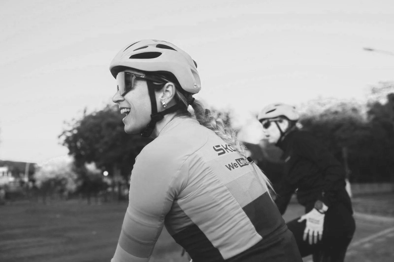Black and white photo of two women wearing helmets and cycling gear, smiling and enjoying a bike ride outdoors.
