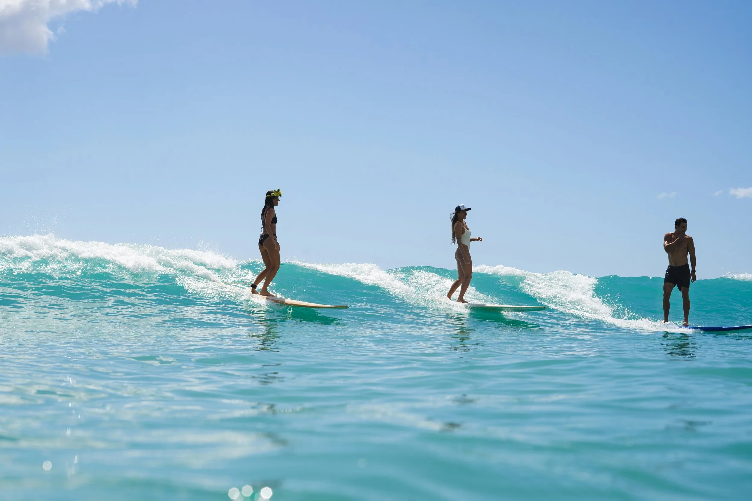 Three people surfing on a clear blue ocean wave, with a light blue sky above.