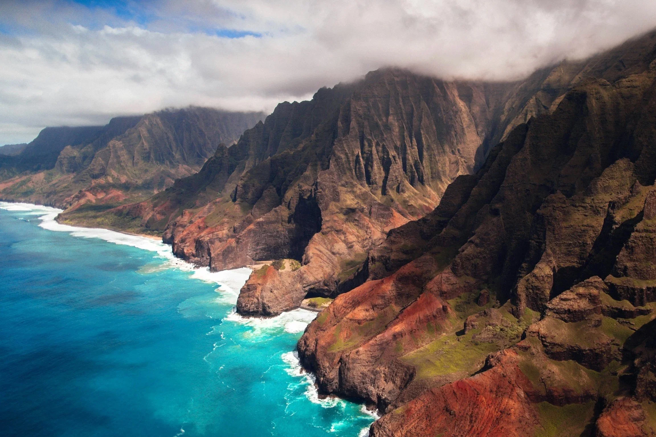 Aerial view of rugged volcanic cliffs along a turquoise ocean coastline with cloud-covered mountains in the background.
