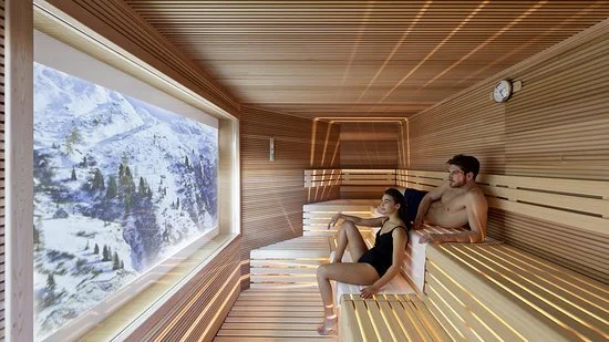 Two people sitting inside a wooden sauna with a large window showing a snowy mountainous landscape outside.