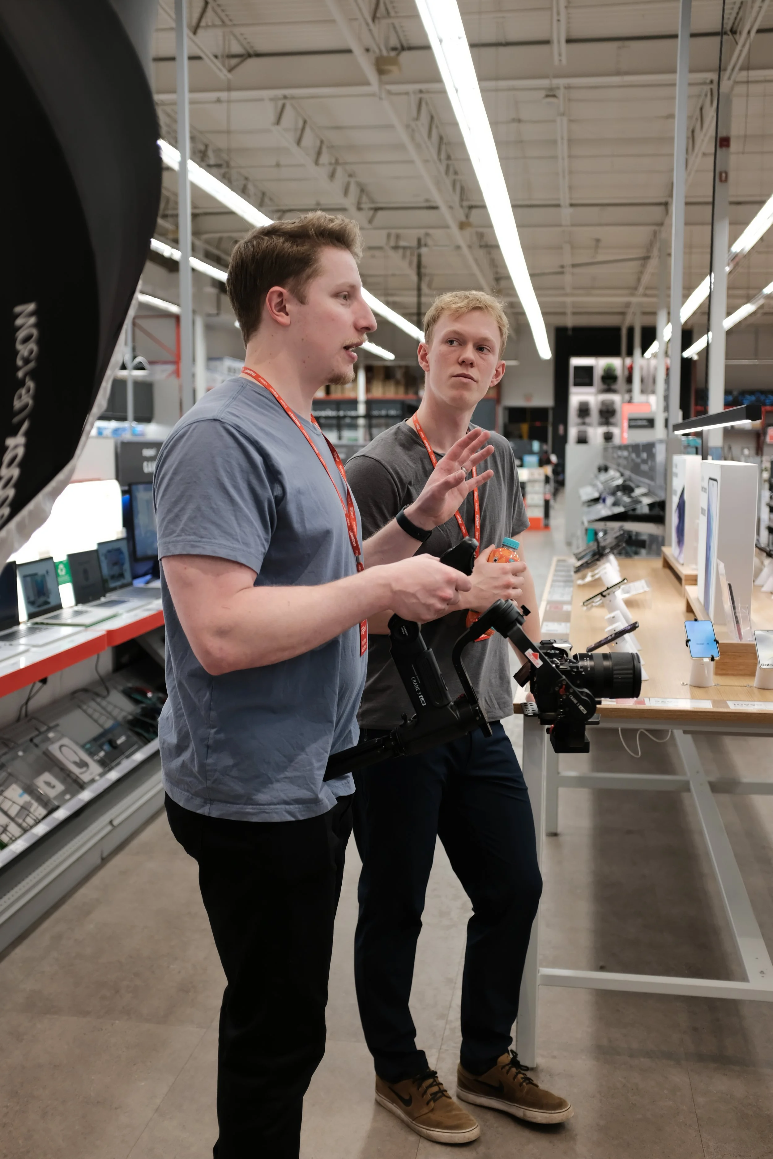 Two men discussing in a tech store, one holding a camera. Laptops and mobile devices displayed on a table.
