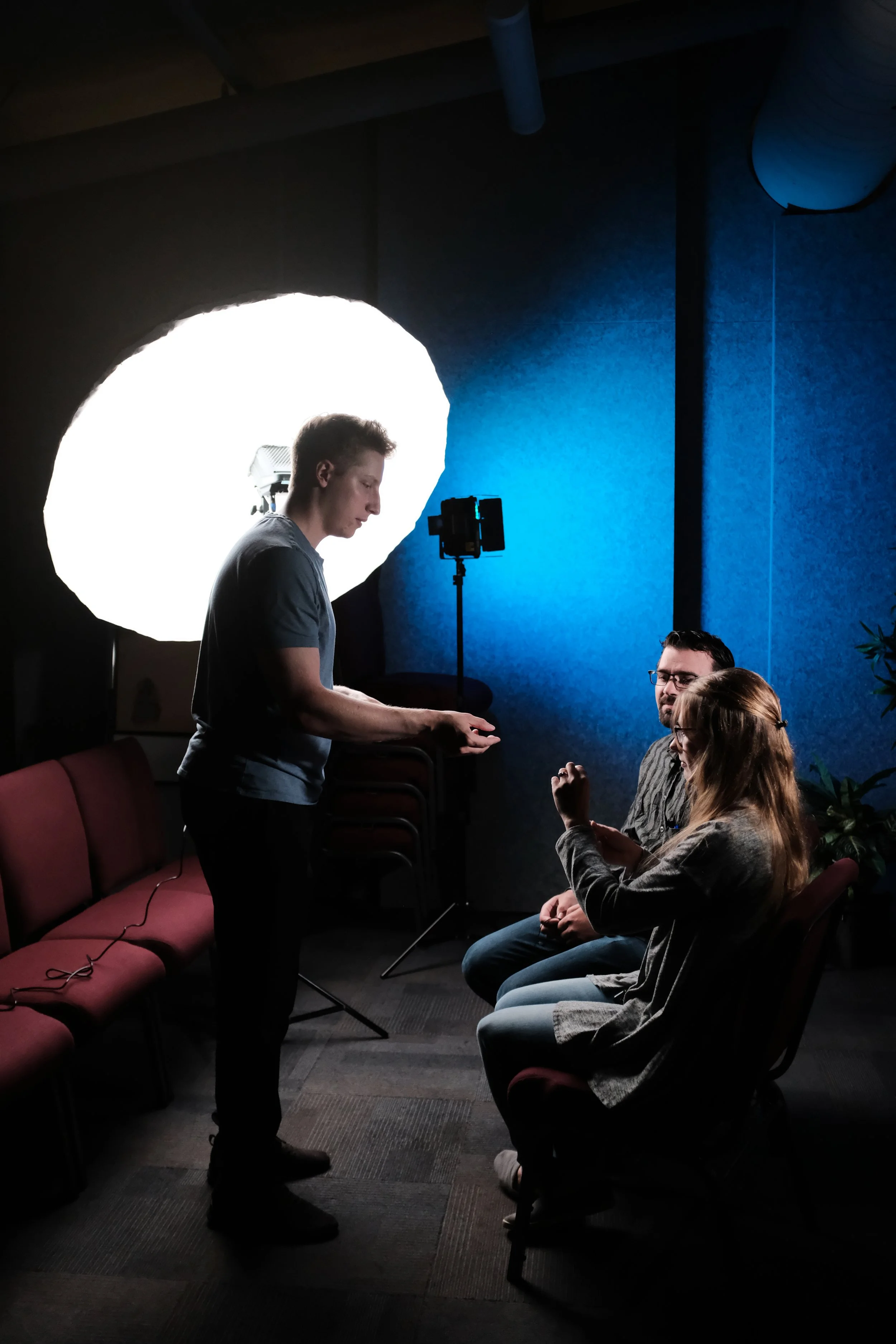 Three people in a dimly lit room with studio lighting equipment, one standing and two sitting discussing something.