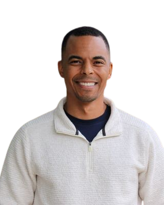 Smiling man wearing a light-colored zip-up sweater with a dark shirt underneath, standing against a plain background.