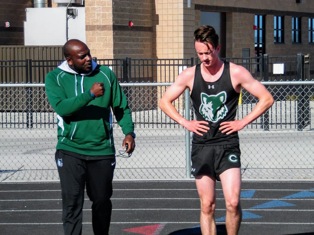 A male track athlete wearing a black and green uniform with a lion logo, standing on a track with hands on hips, looking down, and another man in a green jacket talking to him.