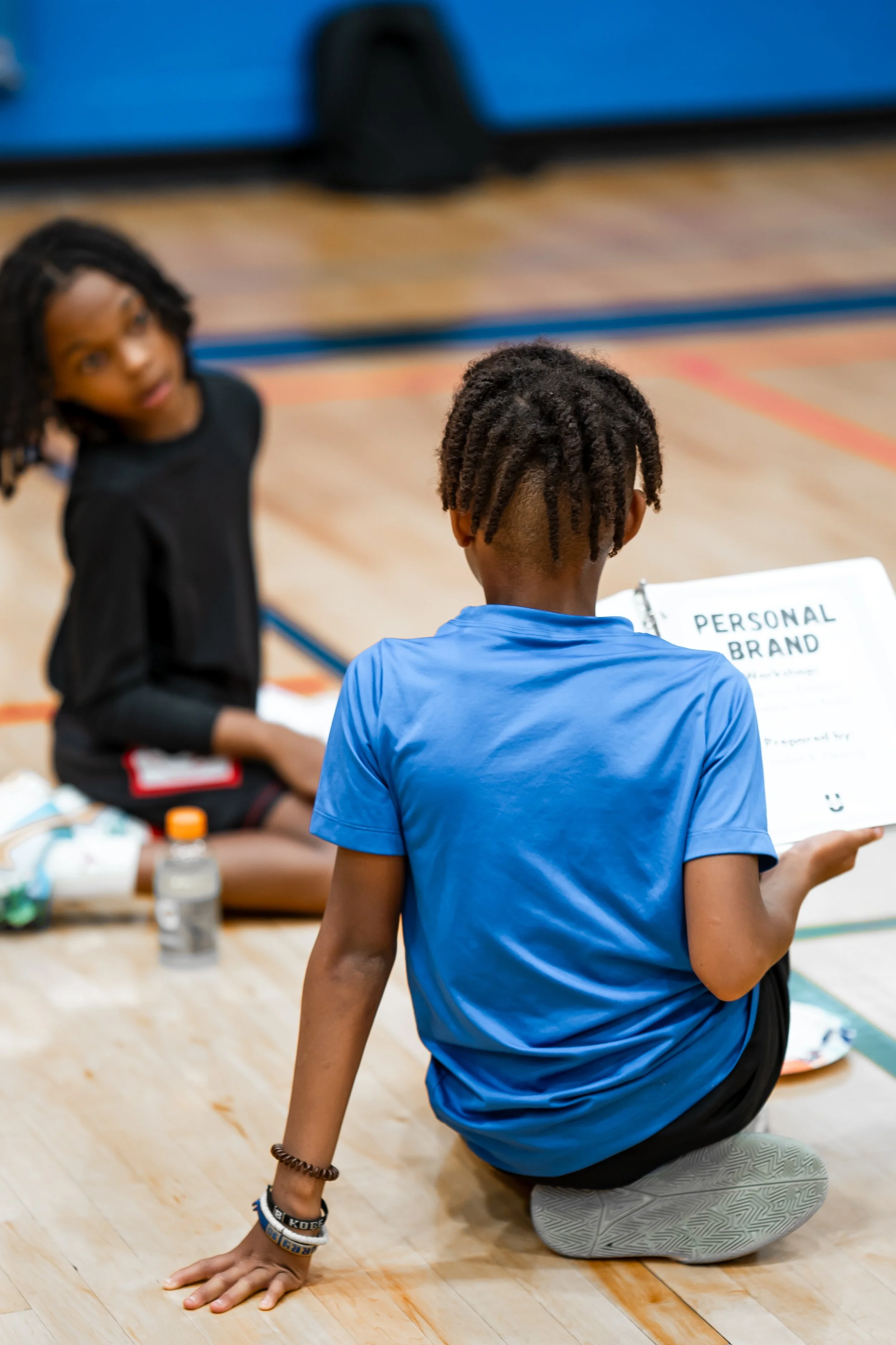 A person in a blue shirt and gray sneakers is kneeling on a gym floor, facing away from the camera, with one hand on the ground. In the background, a woman in black clothing and multiple bracelets is sitting on the gym floor, holding a clipboard, with a bottle of water nearby.