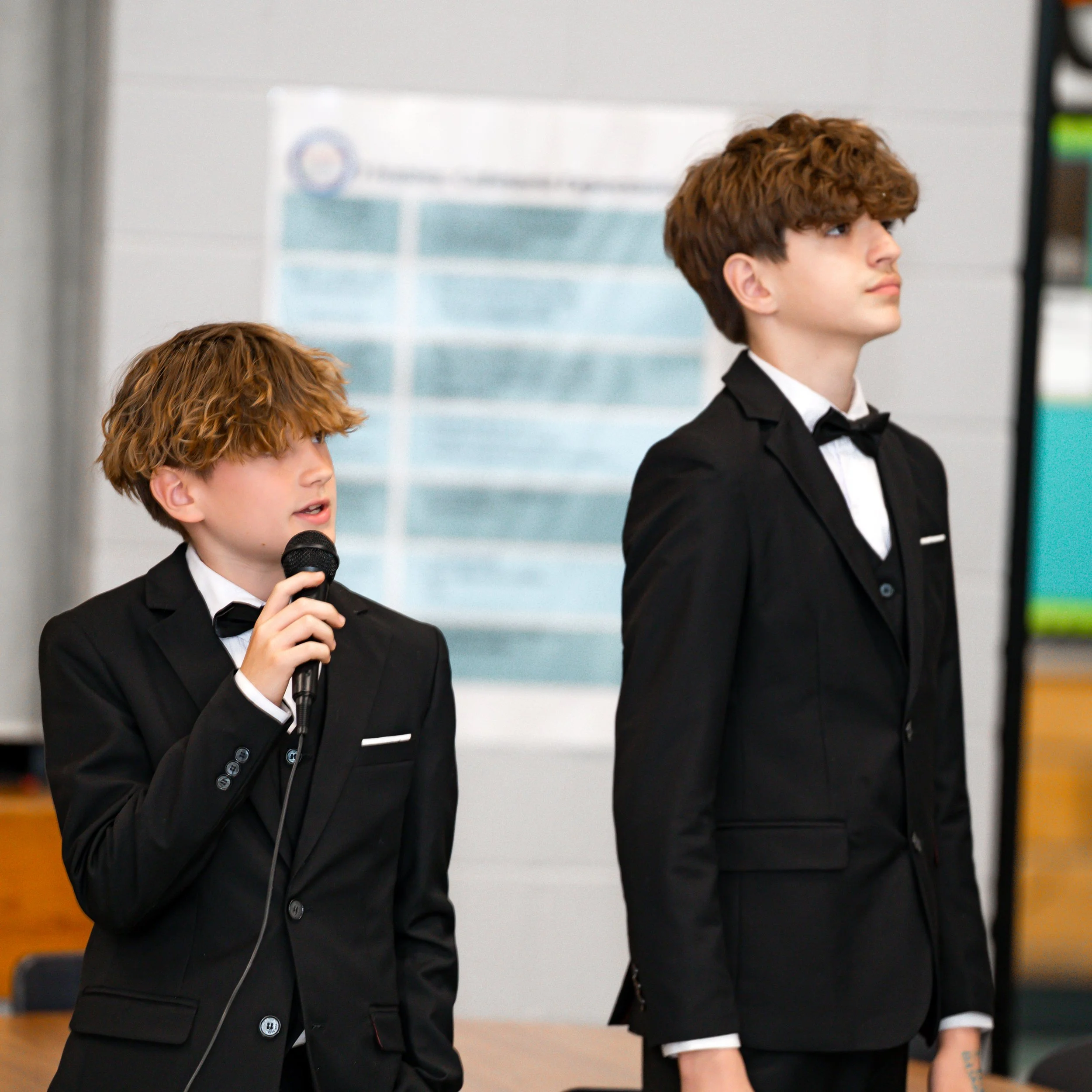 Two young boys dressed in black tuxedos with white shirts and black bow ties are attending a formal event. The boy in the foreground has curly red hair and is holding a microphone, while the boy in the background has straight brown hair and is standing with a serious expression. They appear to be on stage or in a presentation setting.