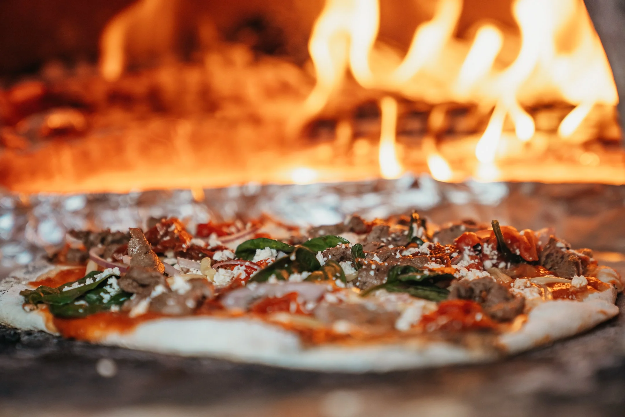 Pizza cooking in a wood-fired oven with flames in the background.