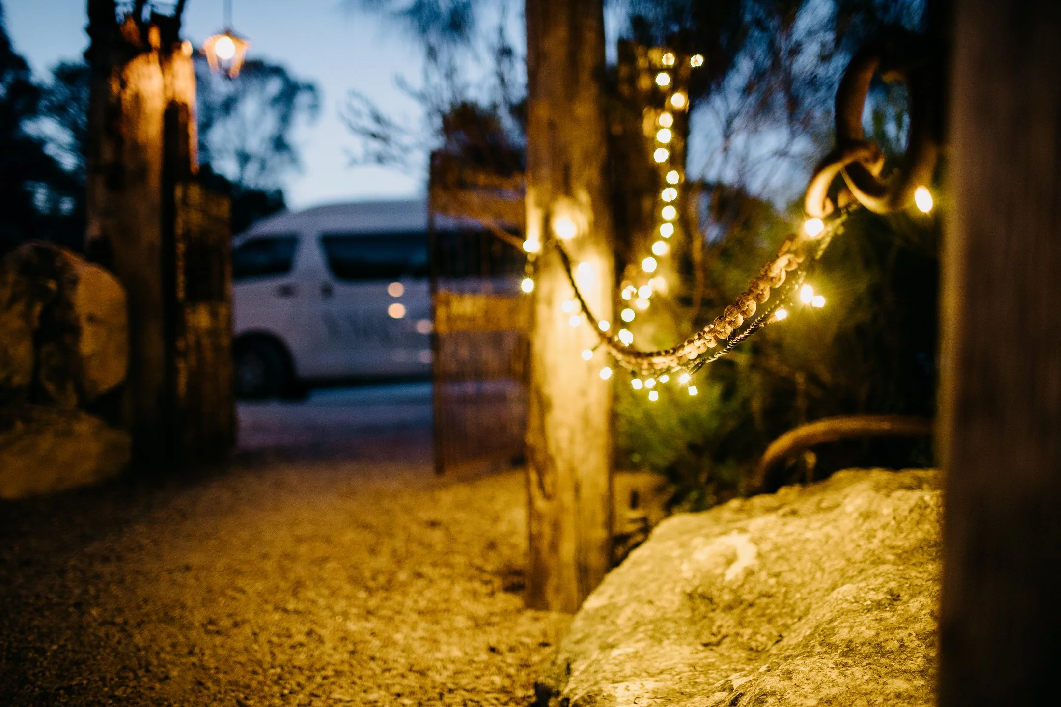 A wooden fence with string lights hanging on it, illuminated in the evening or dusk, with a blurred background of a white van and trees.