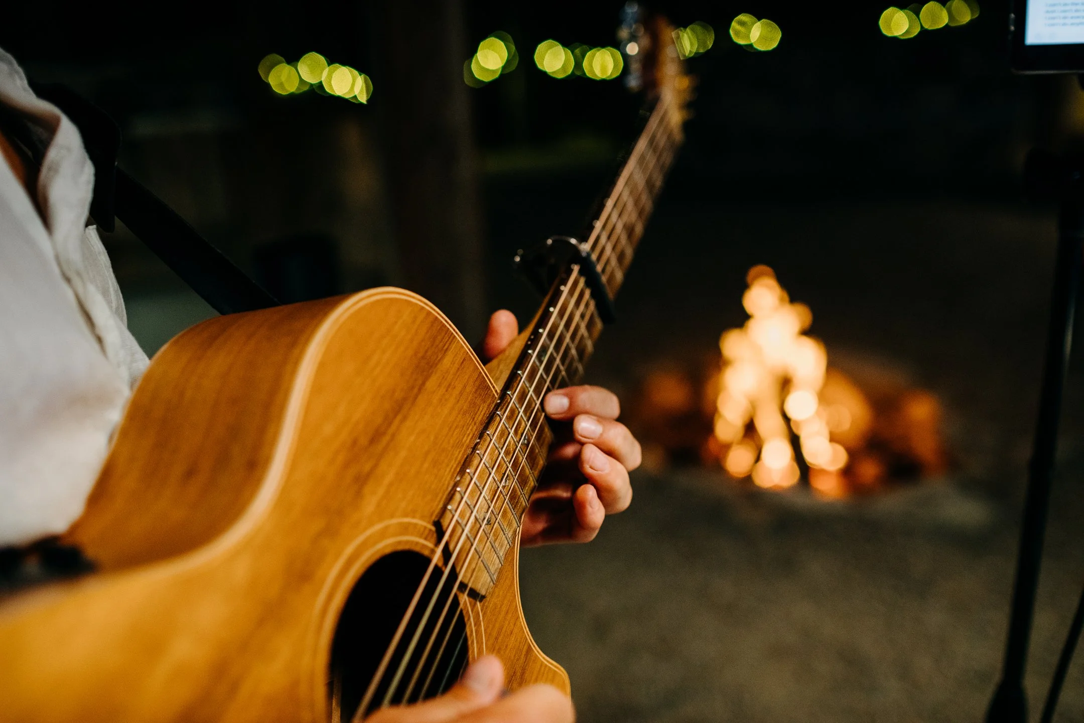 Person playing an acoustic guitar outdoors at night with a campfire and string lights in the background.