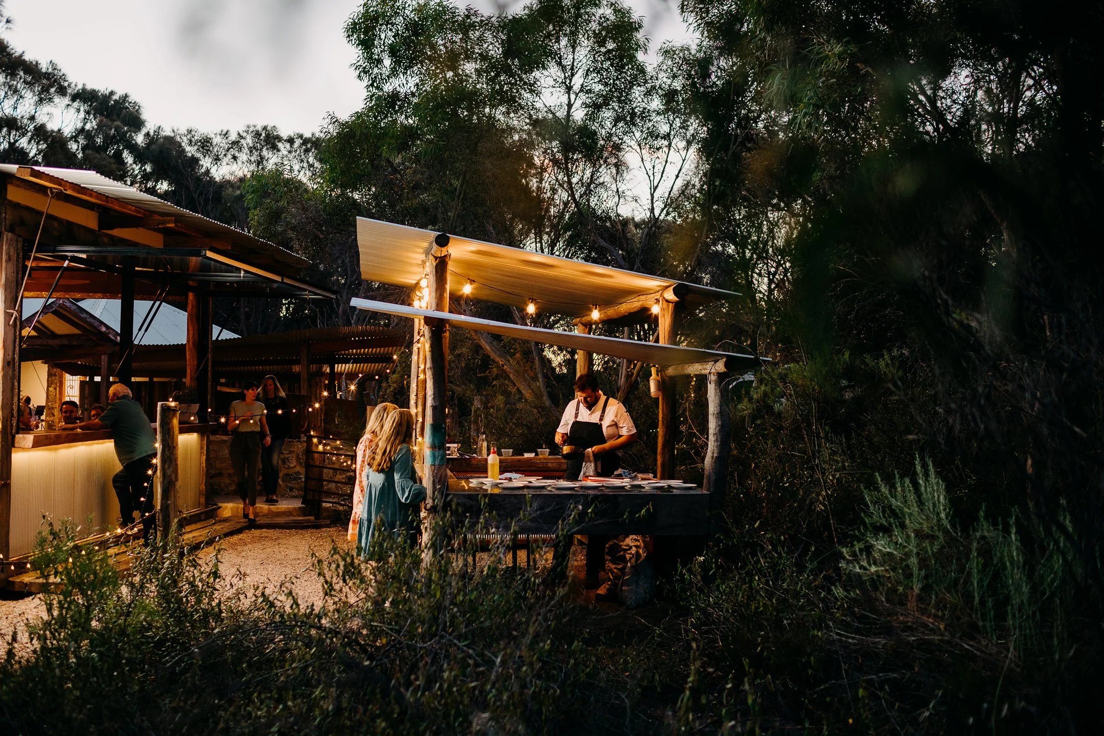 Outdoor dining area at a restaurant during sunset with string lights, people ordering and chatting, and a chef preparing food at a counter.