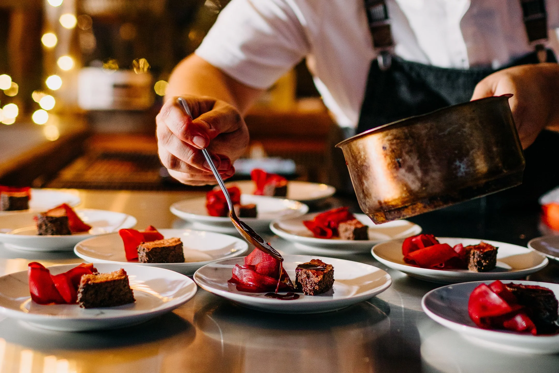 A chef is pouring chocolate sauce onto small slices of cake on white plates, garnished with red edible decorations, in a restaurant kitchen with warm lighting.