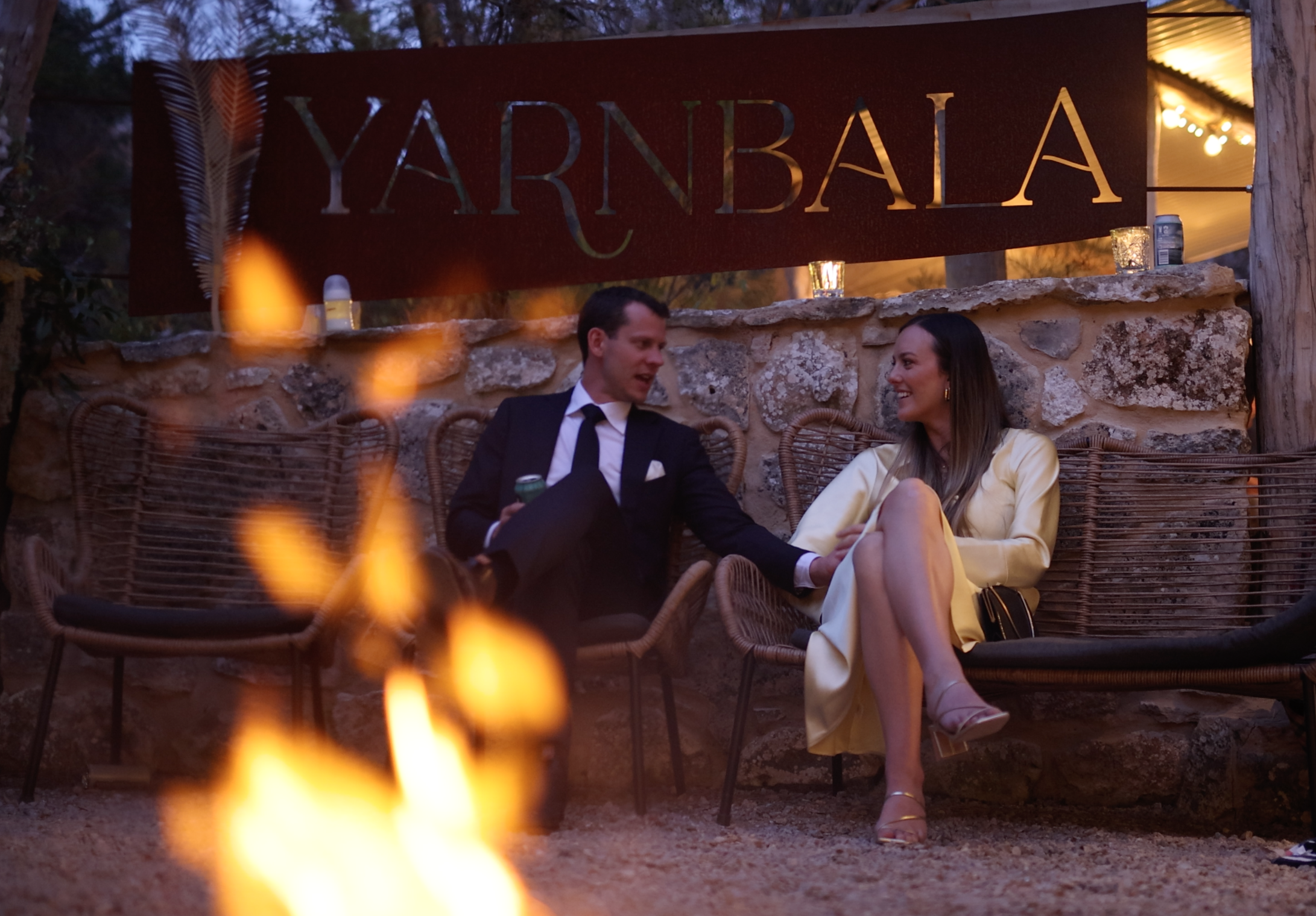 A man and woman sitting on wicker chairs outdoors at night, smiling and talking. A sign behind them reads "YARNBALA." There is a fire in the foreground and string lights overhead, creating a cozy atmosphere.