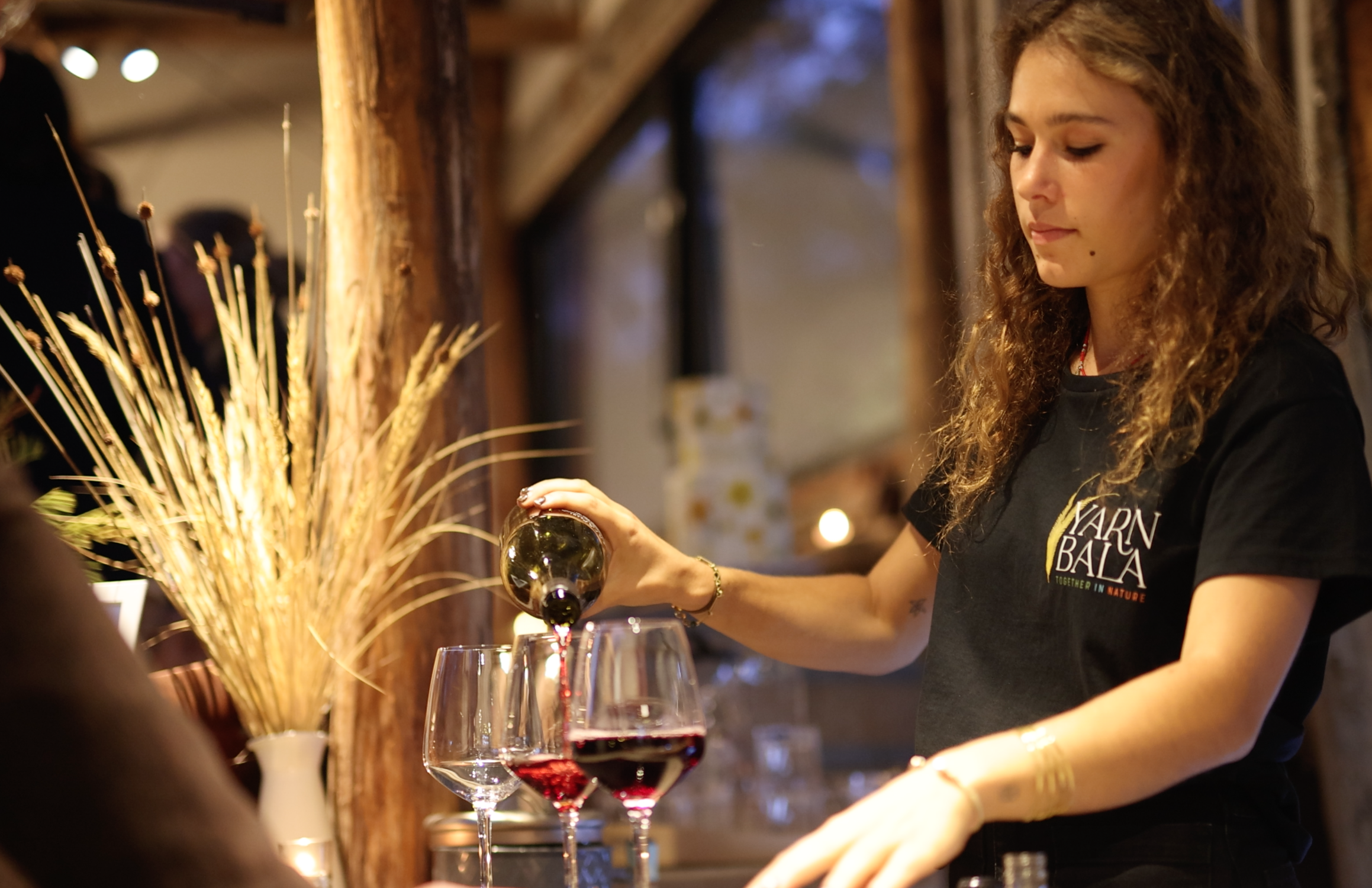 A young woman with curly hair pouring red wine into a glass at a rustic indoor event.
