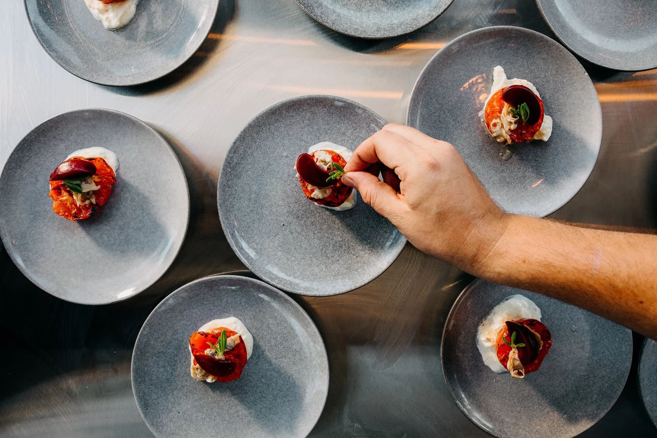 A chef hands garnishes small gray plates with a roasted red pepper, cheese, and green herb.