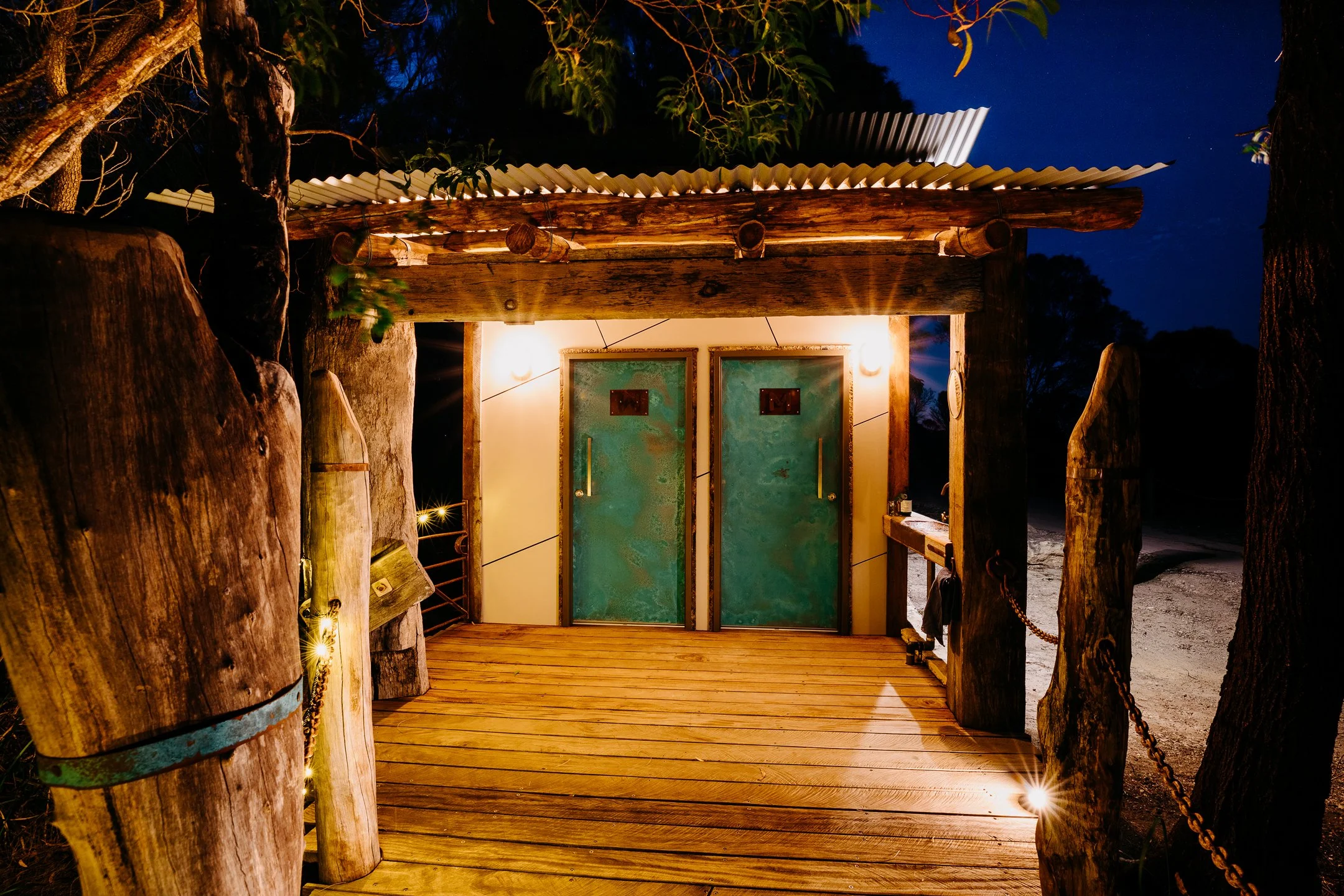 Wooden entrance with teal doors illuminated by warm lights, surrounded by trees and rustic wooden posts, at night.