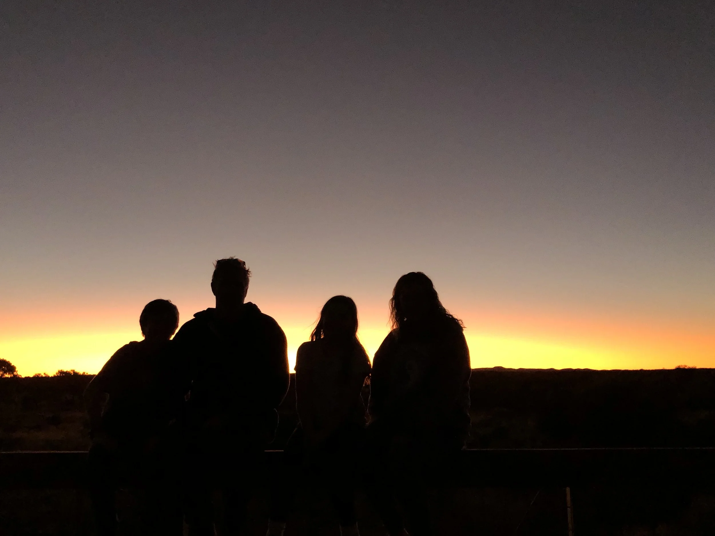 Four silhouetted people sitting on a ledge watching a sunset or sunrise in a desert landscape.