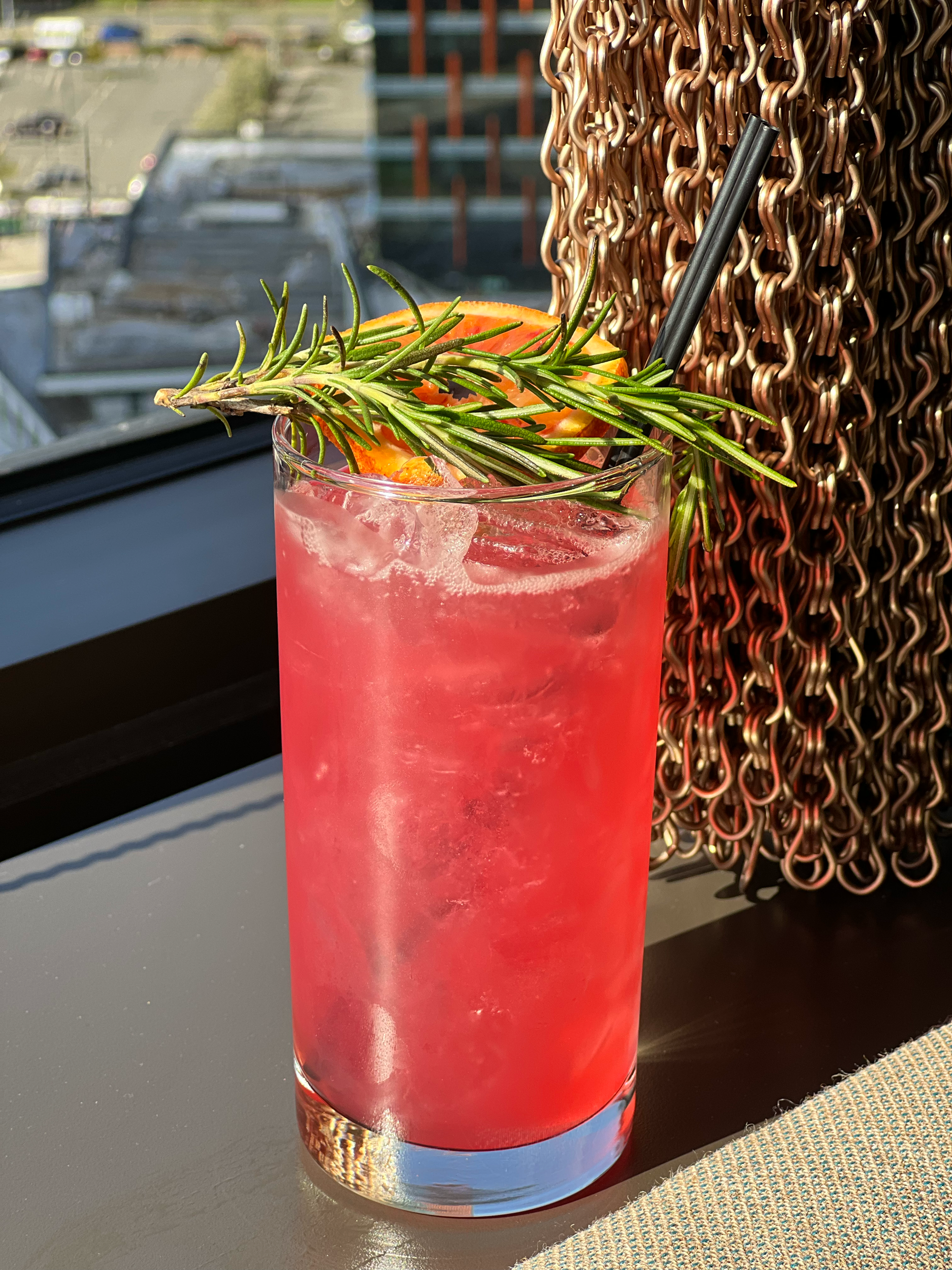 A cold, pink-colored beverage with ice in a clear glass, garnished with a sprig of rosemary and a slice of orange, sitting on a table near a woven placemat.