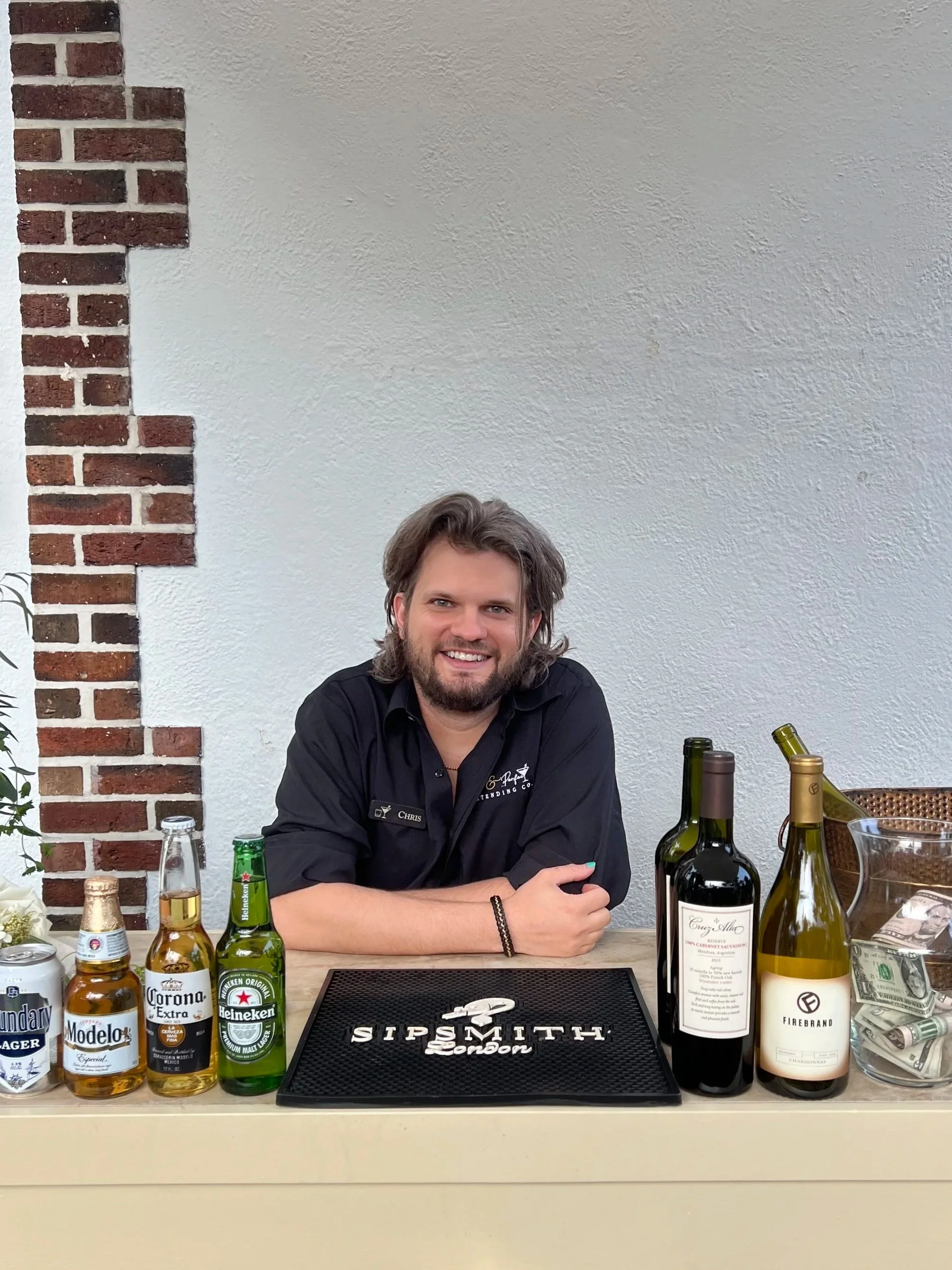 A smiling man behind a bar counter with various wine bottles, beer, and a tip jar, in a modern bar with brick wall decor.